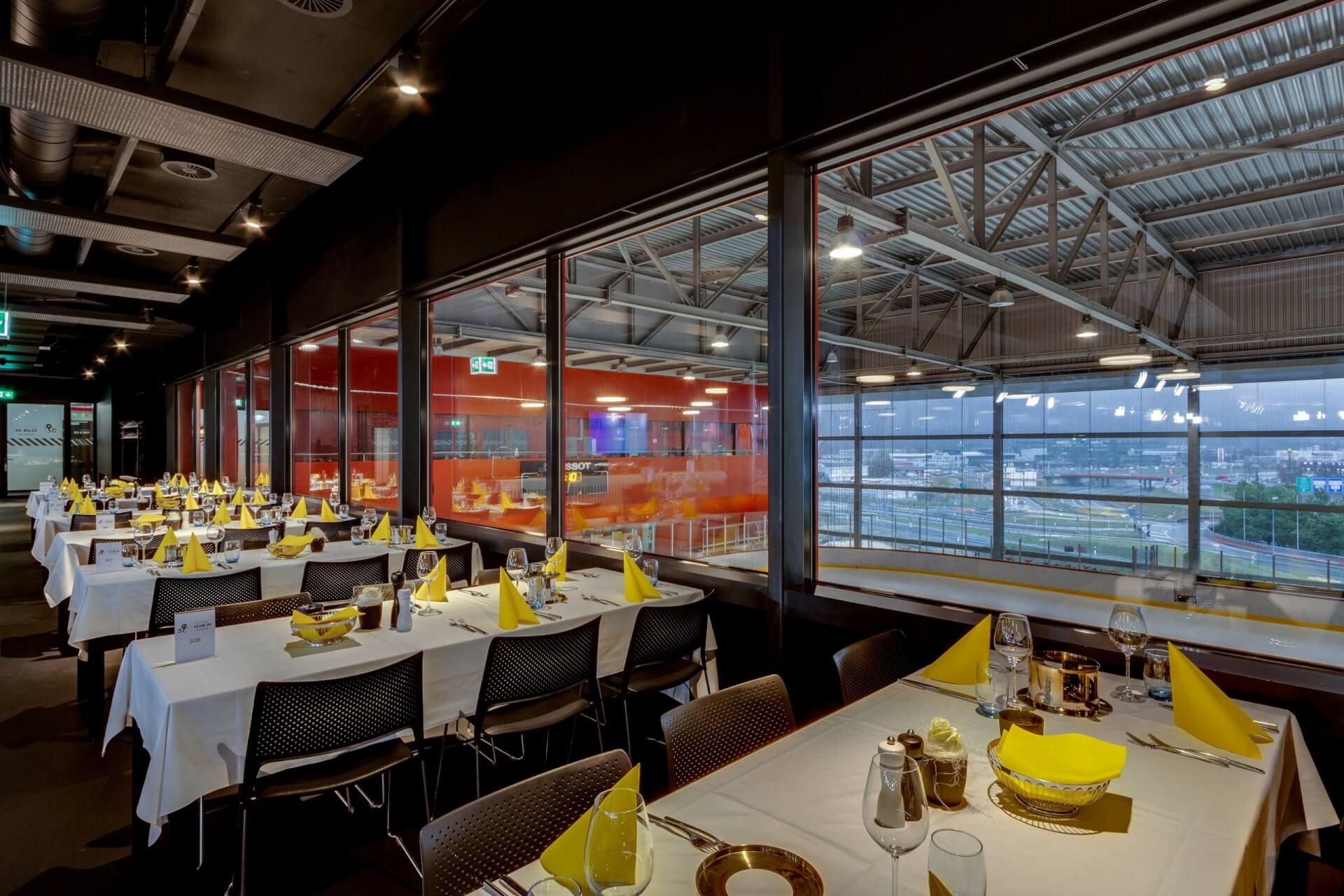 Restaurant mit gedeckten Tischen für die Gäste, mit Blick auf eine Indoor-Gokartbahn durch große Glasfenster.