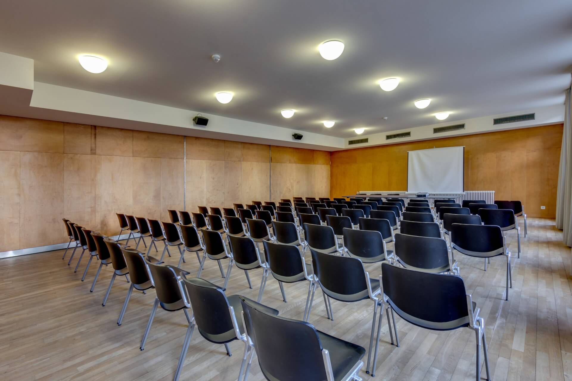 In einem gut beleuchteten Konferenz- oder Vortragsraum mit Holzwänden und Holzfußboden stehen leere schwarze Stühle vor einer Projektionsfläche.