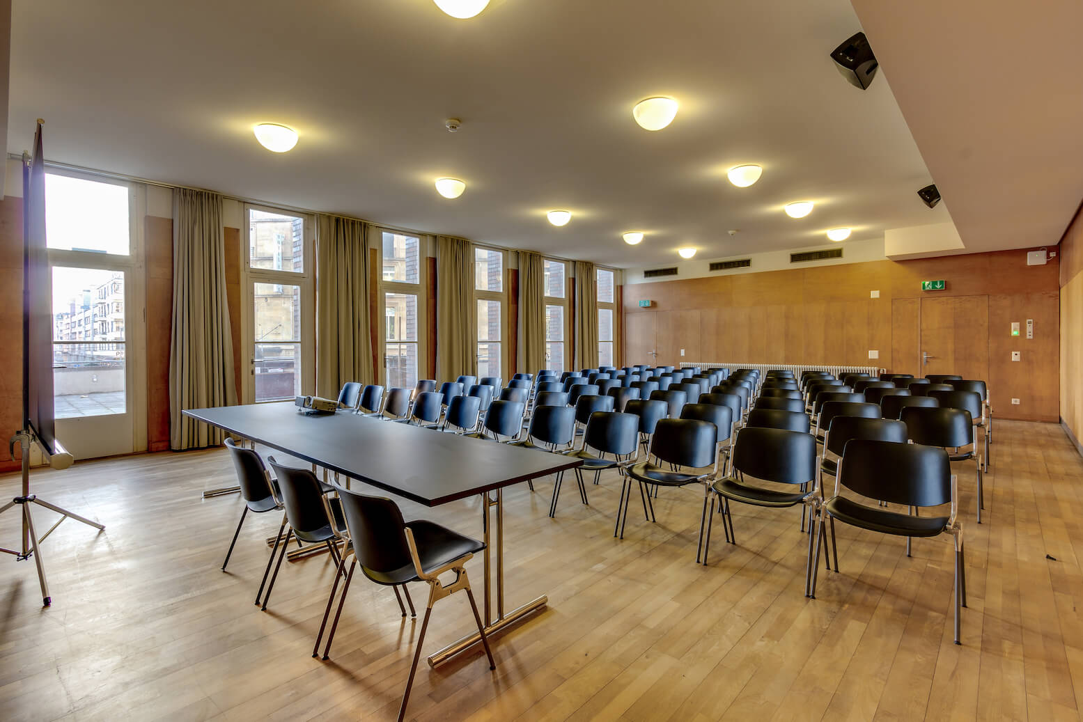Ein Konferenzraum mit Reihen leerer schwarzer Stühle vor einem langen Tisch an der Stirnseite, großen Fenstern an einer Seite und Holzfußboden.
