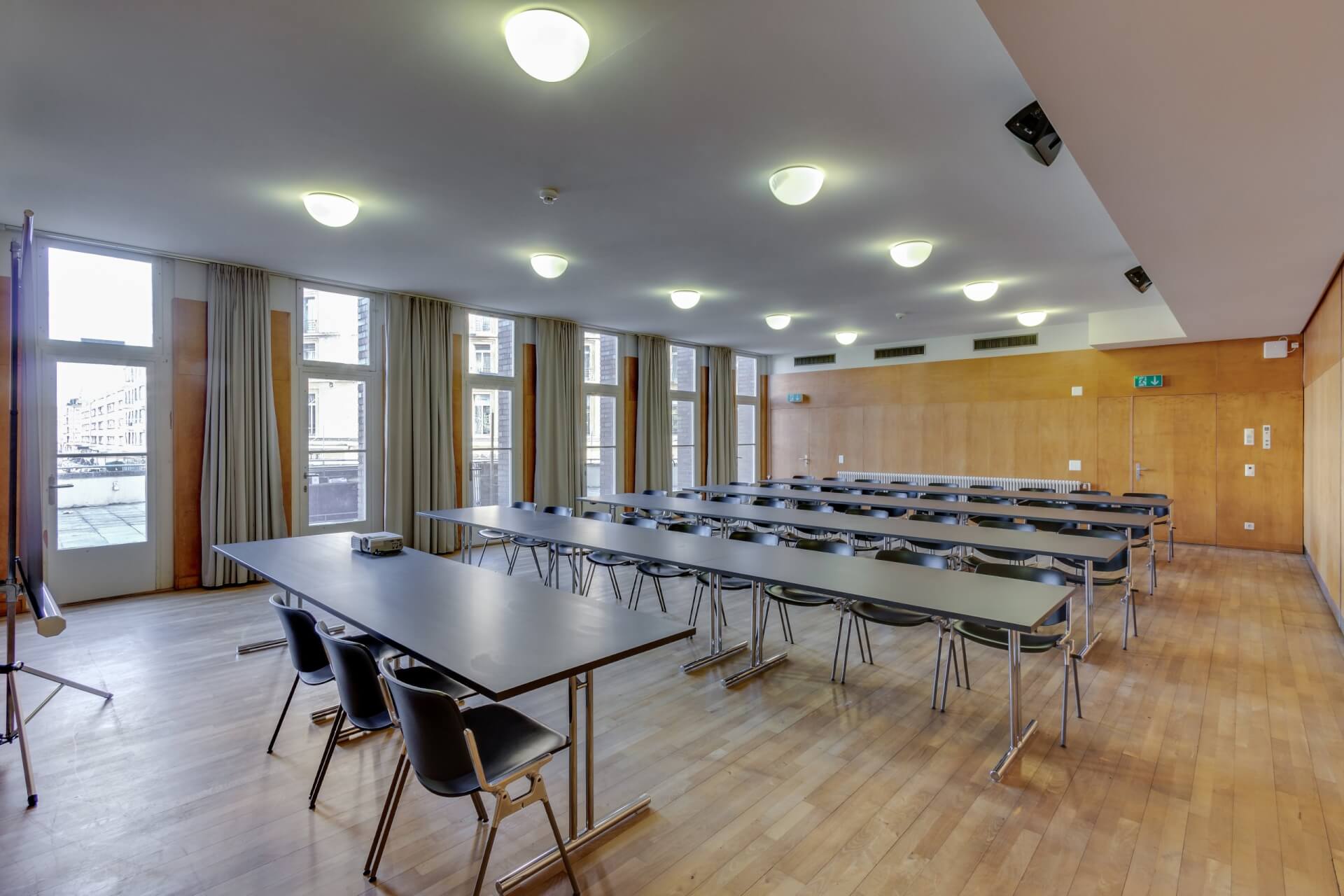 Ein Klassenzimmer mit langen Tischen und schwarzen Stühlen, die in Reihen angeordnet sind, großen Fenstern an einer Seite und holzgetäfelten Wänden. Der Raum ist leer und gut beleuchtet.