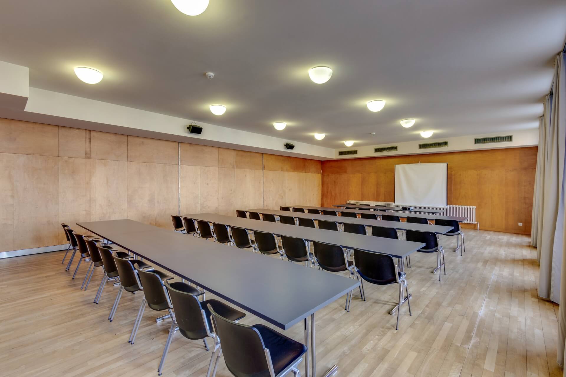 Leeres Klassenzimmer mit drei Reihen langer Tische und schwarzer Stühle vor einer weißen Projektionsfläche auf einem Holzboden.