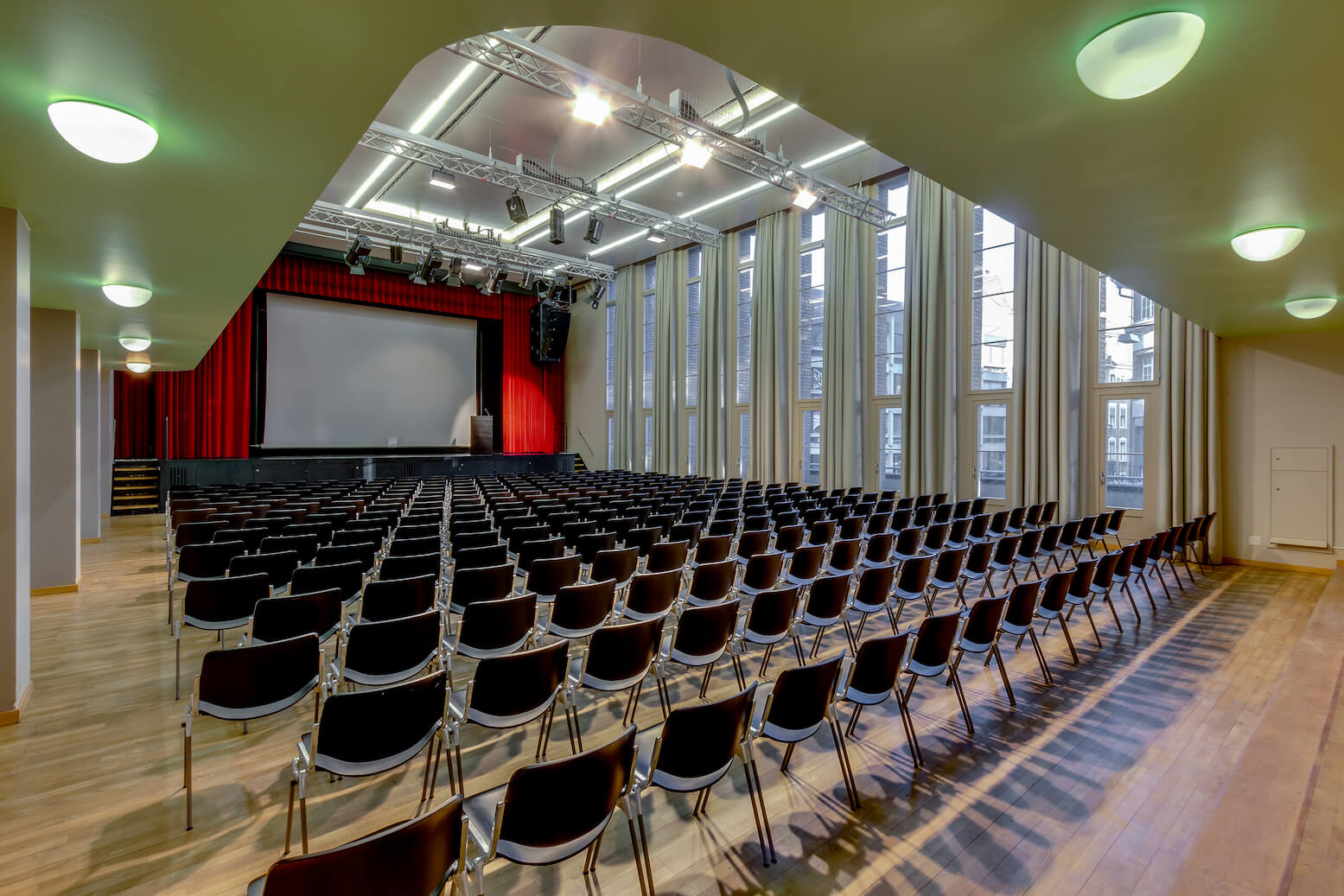 Großer Zuschauerraum mit leeren Stuhlreihen vor einer Bühne mit Leinwand und roten Vorhängen; hohe Fenster auf der rechten Seite und Deckenbeleuchtung erhellen den Raum.