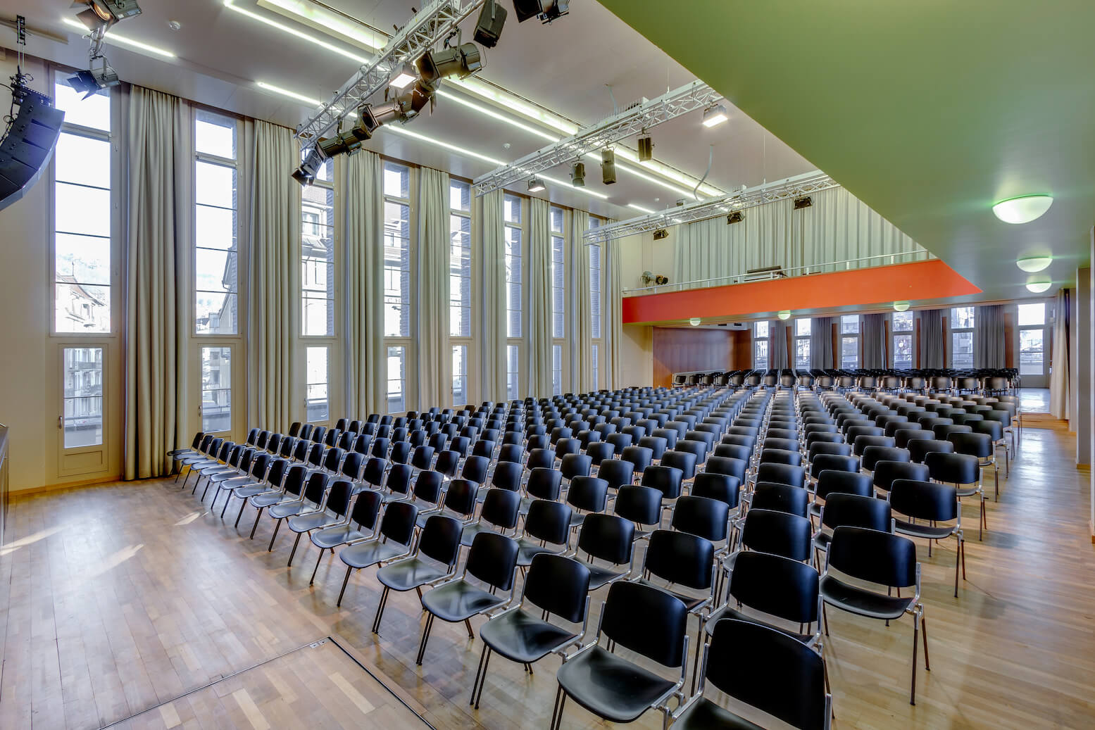 Schwarze Stuhlreihen stehen in einem großen, gut beleuchteten Hörsaal mit Holzfußboden, hohen Decken und großen Fenstern.