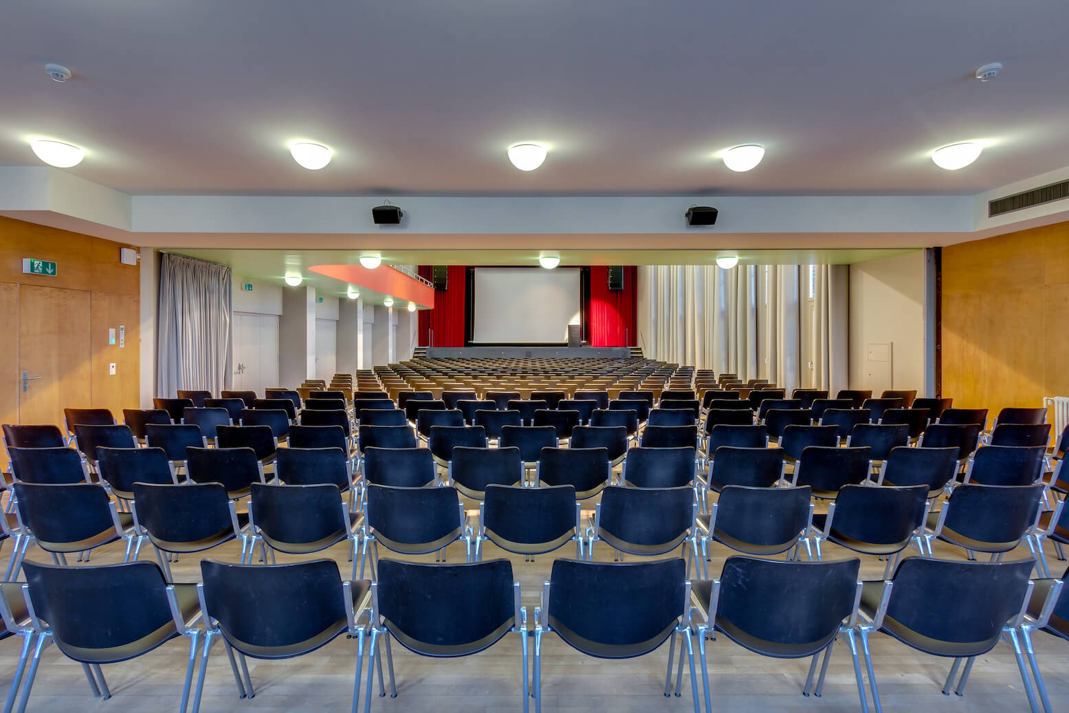 In einem Auditorium sind Reihen leerer schwarzer Stühle vor einer großen Projektionsfläche und einer Bühne mit roten Vorhängen aufgestellt.
