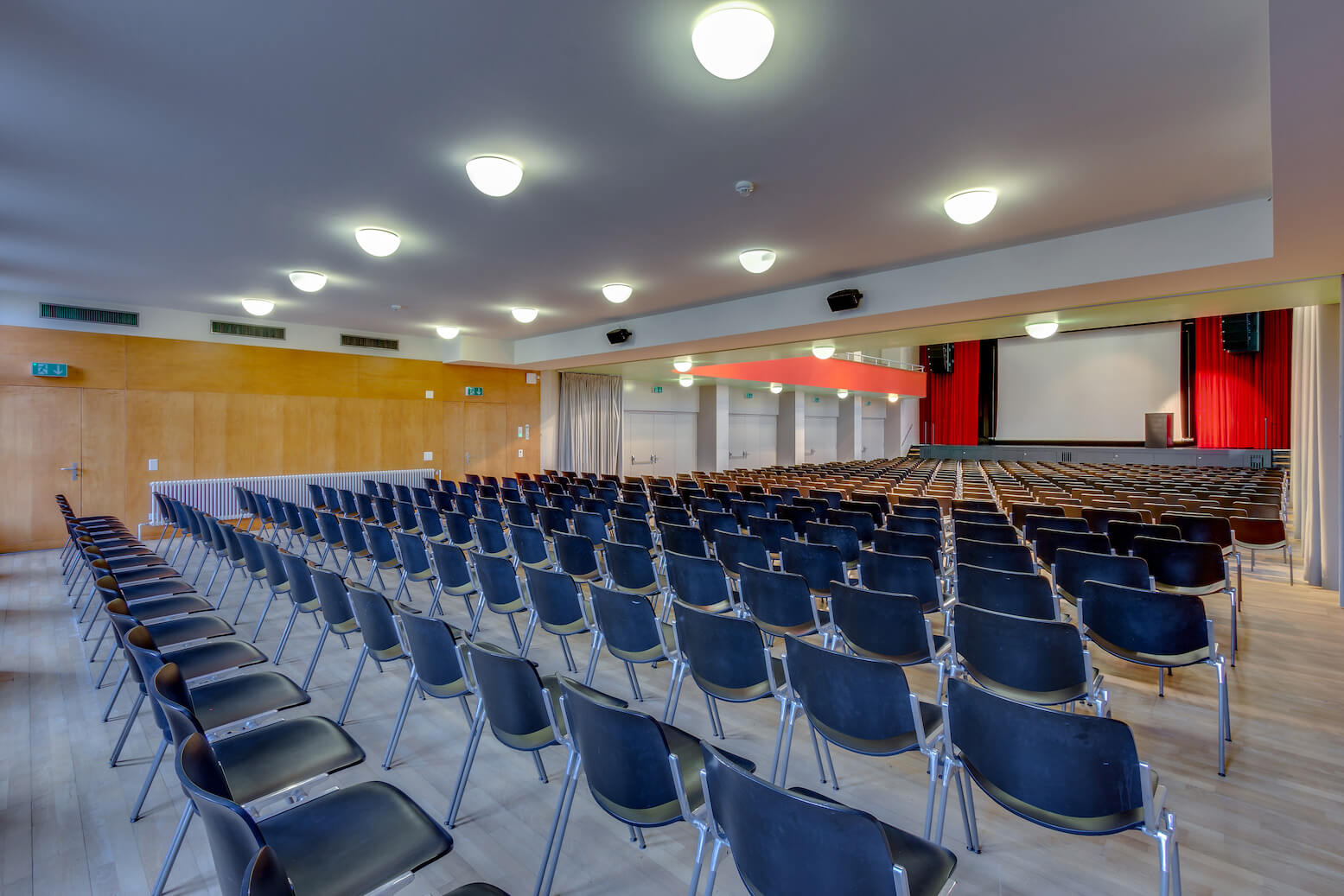 Großer leerer Saal mit Reihen schwarzer Stühle vor einer Bühne mit Projektionsfläche und roten Vorhängen, beleuchtet von Deckenleuchten.