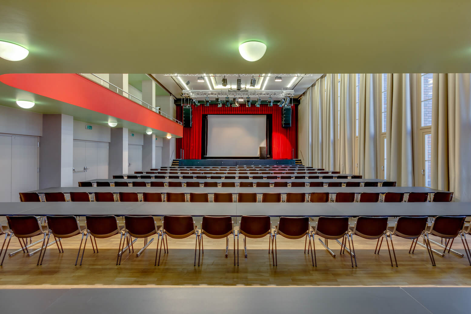 Ein modernes Auditorium mit Reihen leerer Stühle und Tische vor einer Bühne mit großer Leinwand und roten Vorhängen, unter hellen Deckenleuchten.