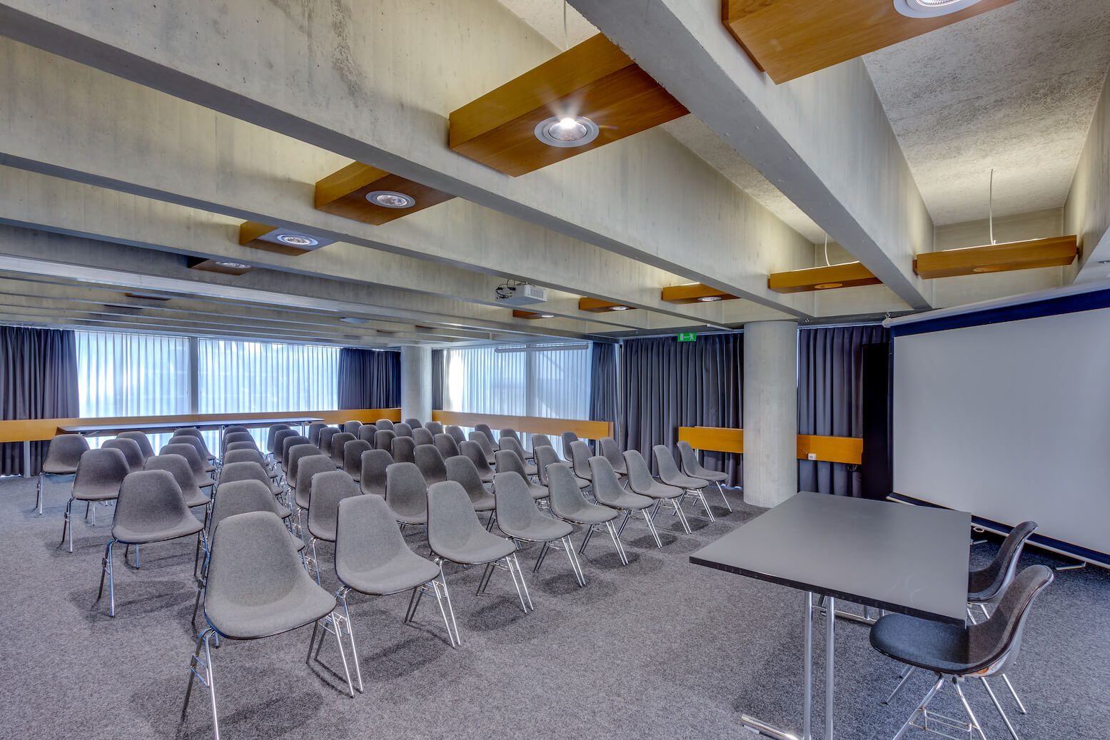 Konferenzraum mit Reihen leerer grauer Stühle vor einer Projektionsfläche, Tischen und bodentiefen Fenstern mit geschlossenen Vorhängen.
