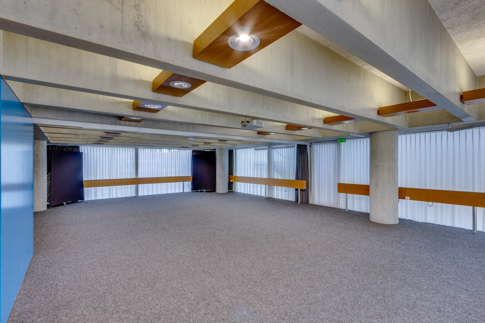 Ein großer, leerer Raum mit Teppichboden, sichtbaren Betonbalken, Deckendetails aus Holz und Metall und raumhohen Fenstern mit vertikalen Jalousien.