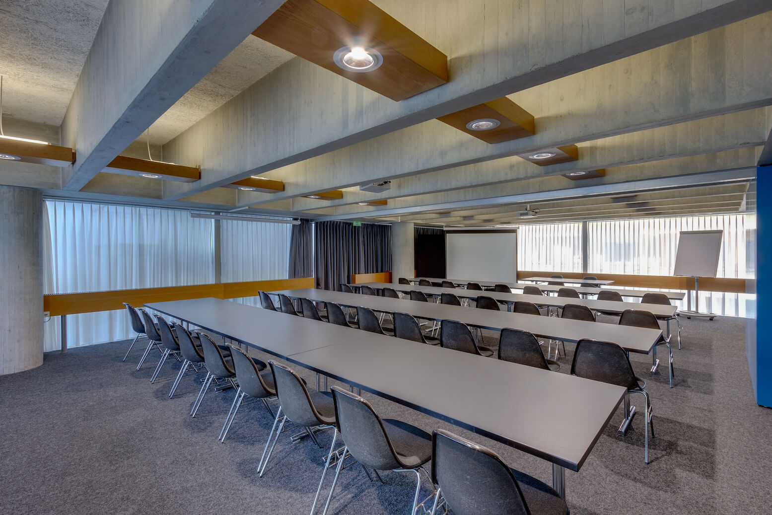 Ein moderner Konferenzraum mit langen Tischen und schwarzen Stühlen, vor denen ein Projektor und ein Flipchart stehen, beleuchtet von Deckenleuchten.