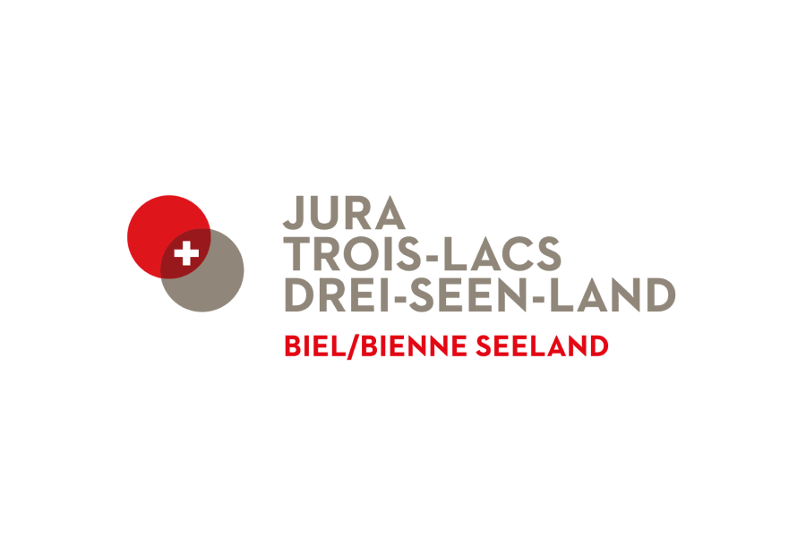 Logo für Jura Trois-Lacs Drei-Seen-Land mit einem roten und grauen Kreis, einem weißen Schweizer Kreuz und Text in Französisch, Deutsch und rotem Englisch darunter.