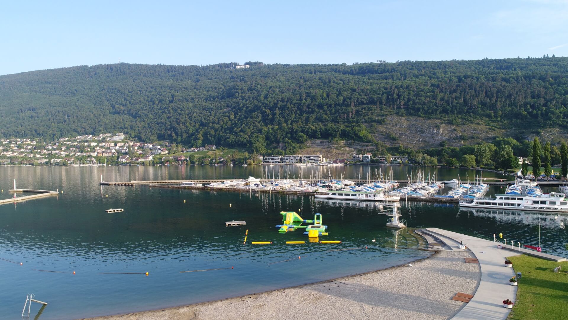 Ein Yachthafen am See mit zahlreichen angedockten Booten, ein schwimmender aufblasbarer Wasserpark, ein Sandstrand und grüne Hügel im Hintergrund.