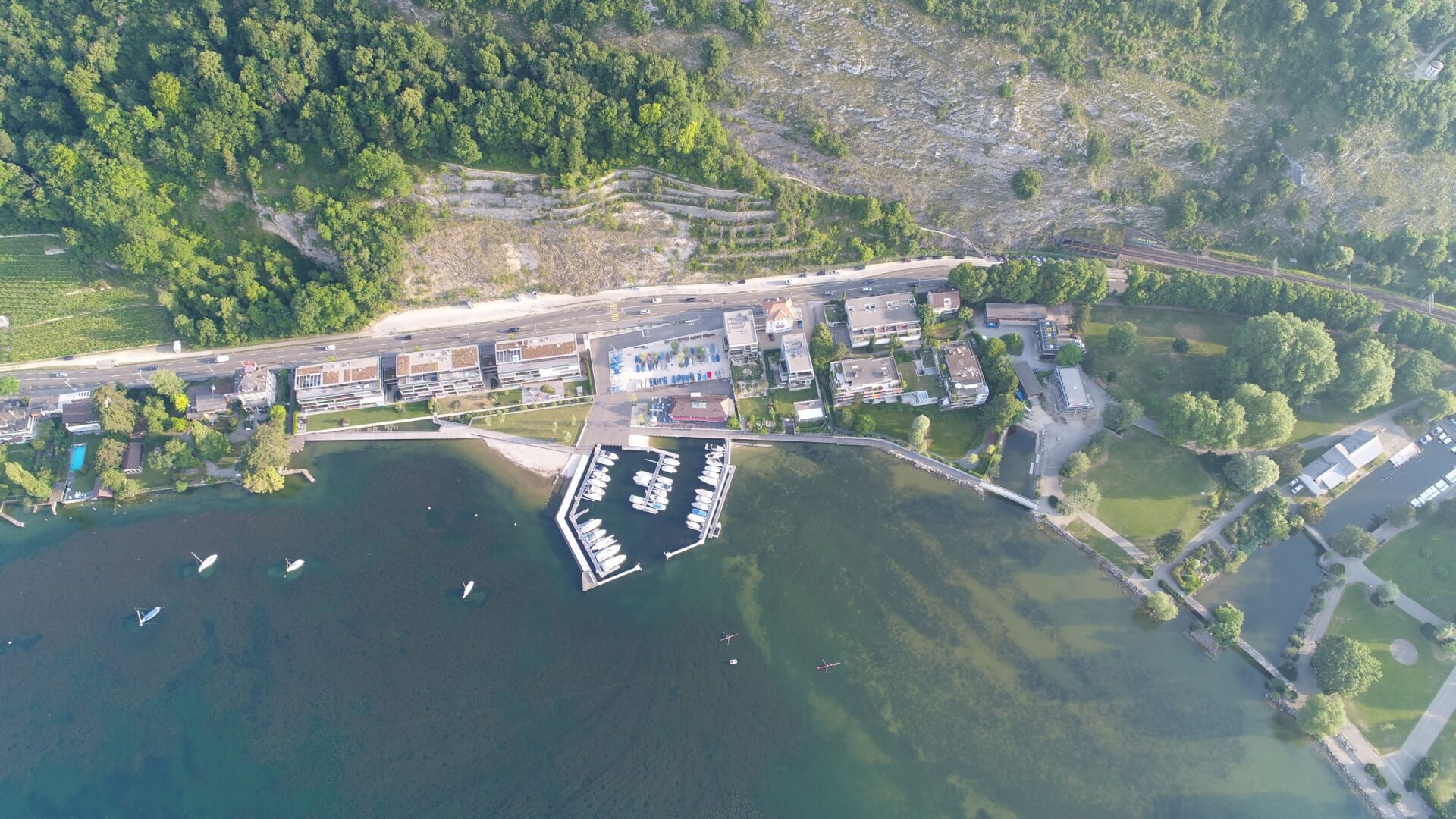 Luftaufnahme einer Küstenstadt mit einem Jachthafen, im Wasser angedockten Booten, Wohngebäuden, einer Hauptstraße und Grünflächen mit Bäumen und Gras.
