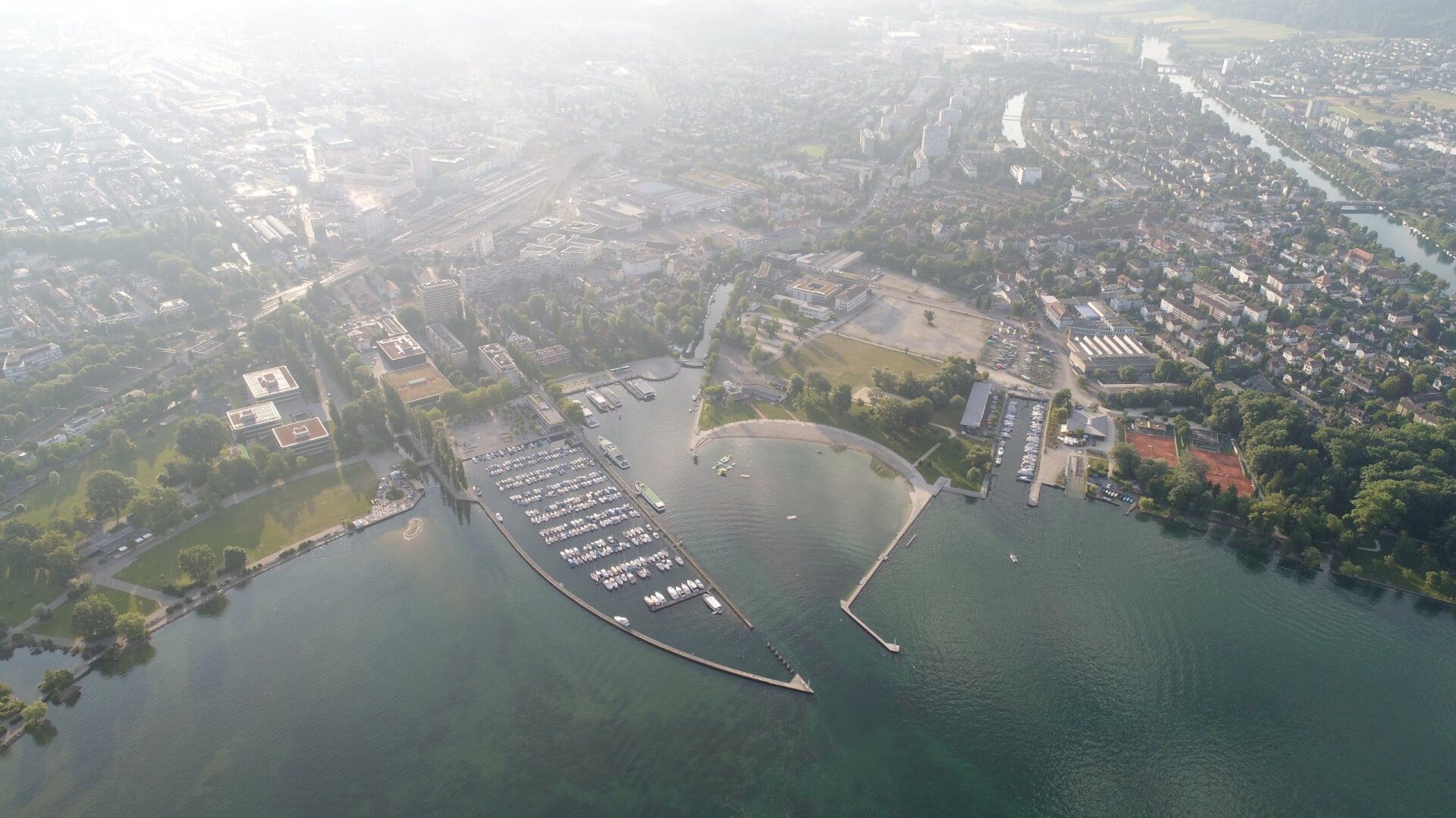 Luftaufnahme eines Jachthafens mit angedockten Booten, angrenzend an eine Stadt und Grünflächen, mit Wasser und Stadtgebieten im Hintergrund.