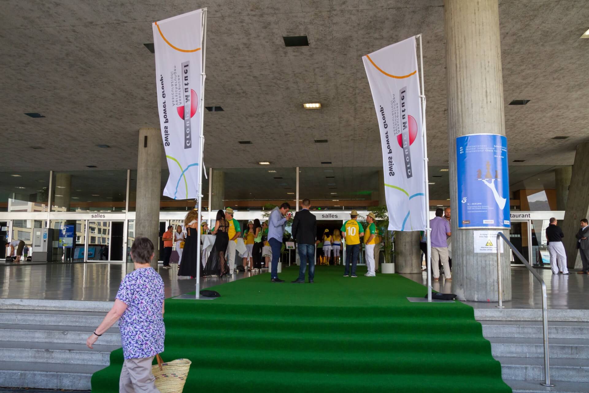 Menschen betreten ein Gebäude mit grünem Teppich und zwei großen Bannern; einige tragen gelbe Trikots, während im Vordergrund eine Frau mit einem Korb vorbeigeht.