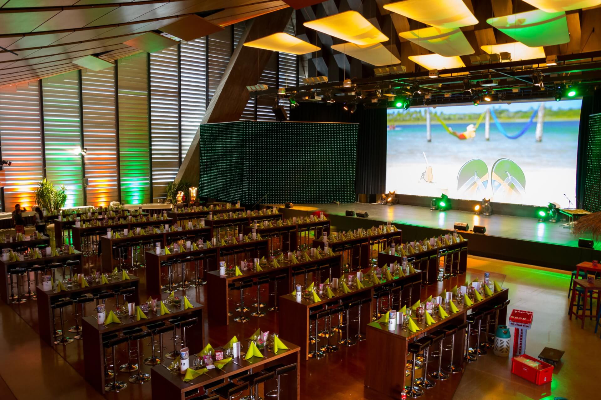Großer Veranstaltungsraum mit Tischreihen mit Partyhüten und Getränken, Bühne im vorderen Bereich mit großer Leinwand und grüner Beleuchtung, darüber moderne Deckenpaneele.