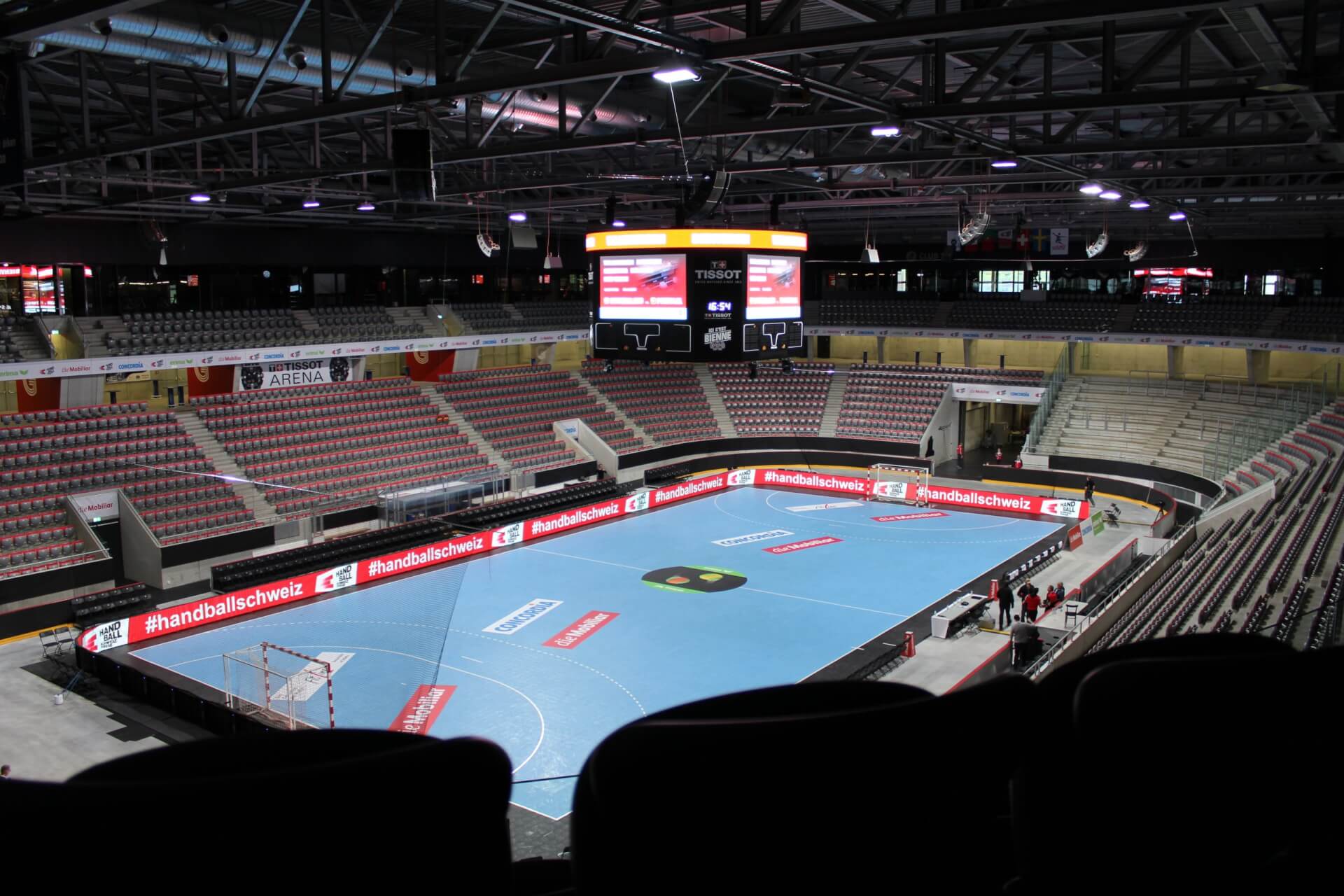 Leere Sporthalle mit blauem Handballfeld, digitalen Werbetafeln und einer großen zentralen Anzeigetafel, auf der Veranstaltungsinformationen angezeigt werden.