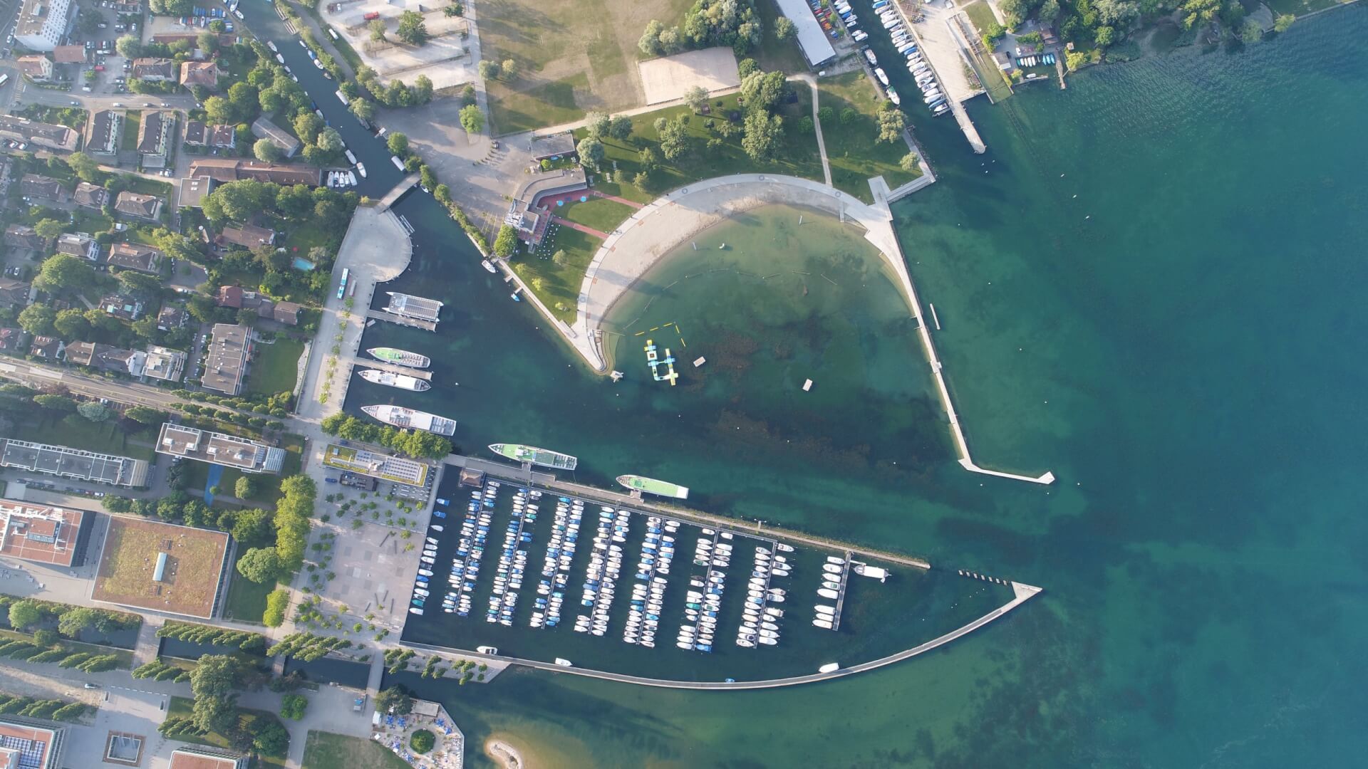 Luftaufnahme eines Jachthafens mit vielen angedockten Booten, geschwungenen Molen und klarem, blaugrünem Wasser neben einem Park und städtischen Gebäuden.