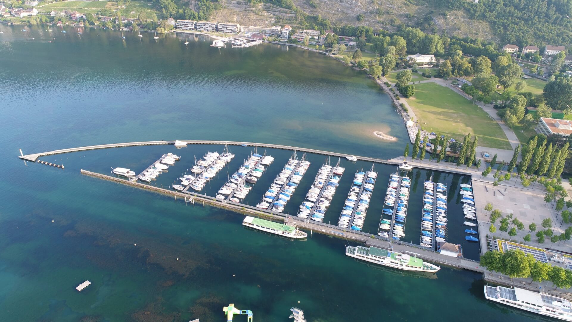 Luftaufnahme eines Jachthafens mit mehreren angedockten Booten, einem geschwungenen Wellenbrecher, einer nahe gelegenen Grünanlage und umliegenden Gebäuden an der Küste.