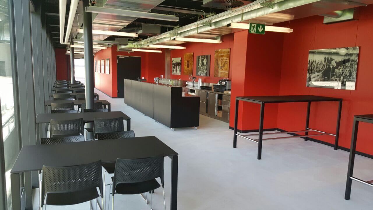 Moderne Cafeteria mit schwarzen Tischen und Stühlen, roten Wänden, industrieller Decke und Küchenbereich im Hintergrund. Gerahmte Fotos hängen an den Wänden.