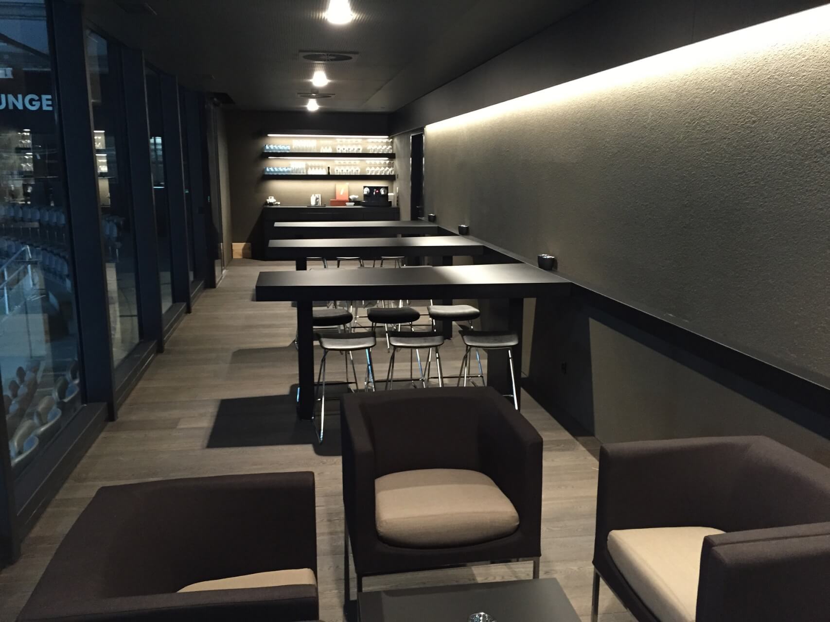 Moderner Lounge-Bereich mit schwarzen Tischen, Stühlen und Sesseln, mit einem Bartresen mit Regalen im Hintergrund und bodentiefen Fenstern an einer Seite.