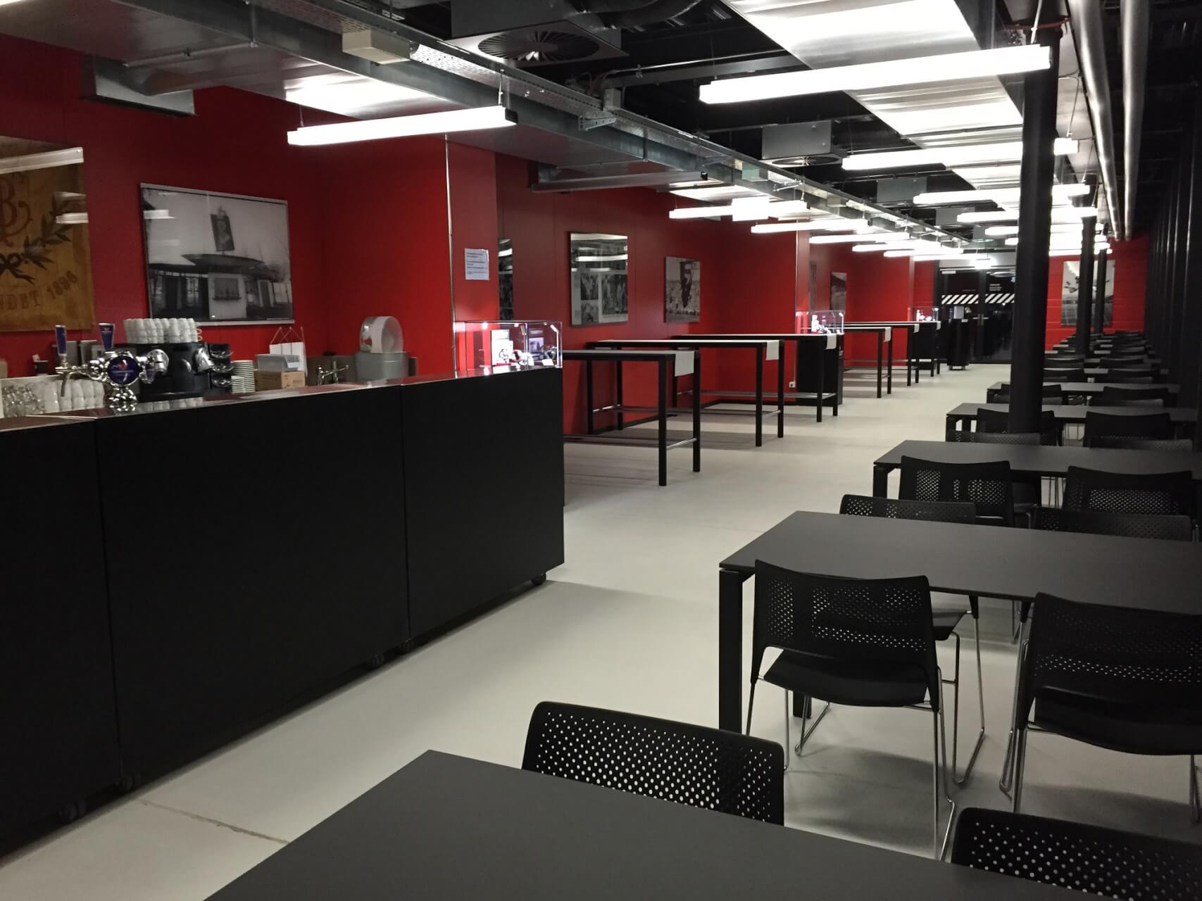 Moderne Cafeteria mit roten Wänden, schwarzen Tischen und Stühlen, hohen Theken auf der linken Seite und Neonbeleuchtung über dem Kopf. Der Raum ist menschenleer.