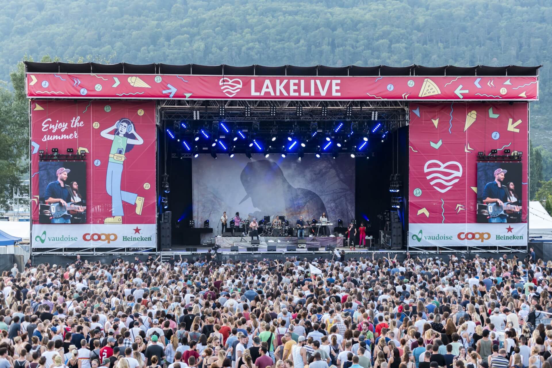 Eine große Menschenmenge steht vor der Freilichtbühne des Lakelive-Festivals. Die Musiker treten unter hellen Bühnenlichtern auf, im Hintergrund ist ein bewaldeter Hügel zu sehen.
