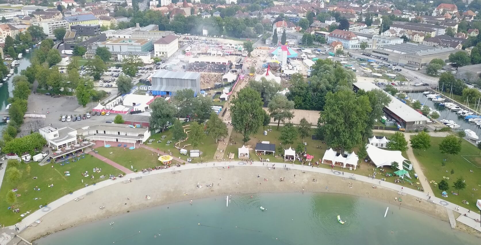 Luftaufnahme eines Strandes am Seeufer mit Menschen, einer Parkwiese, Zelten und einer großen Veranstaltung im Freien mit Menschenmengen und Strukturen im Hintergrund.
