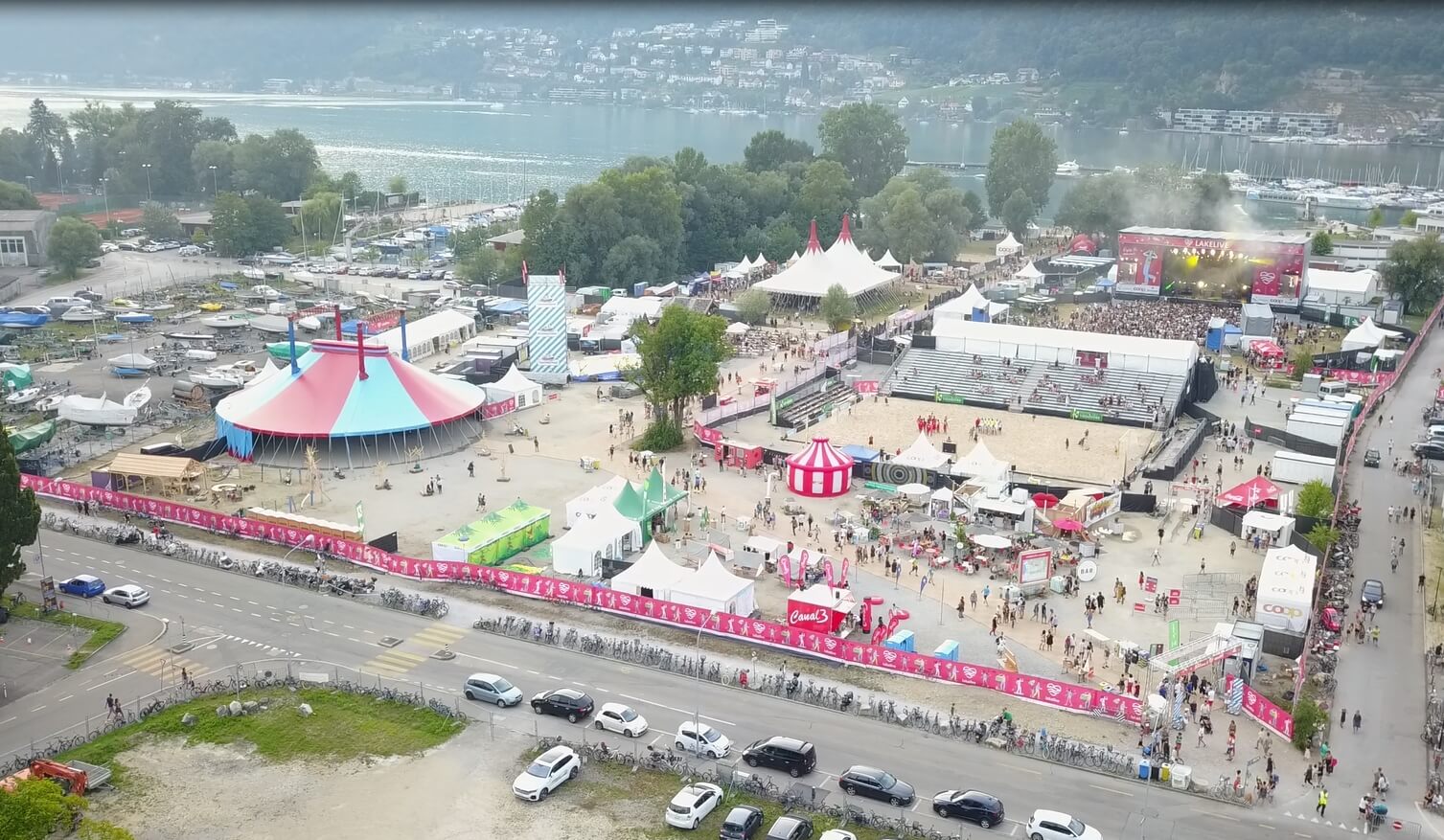 Luftaufnahme eines Festivals am See mit Zelten, Bühnen und Menschenmassen, umgeben von Bäumen und geparkten Autos, mit Hügeln und Wasser im Hintergrund.