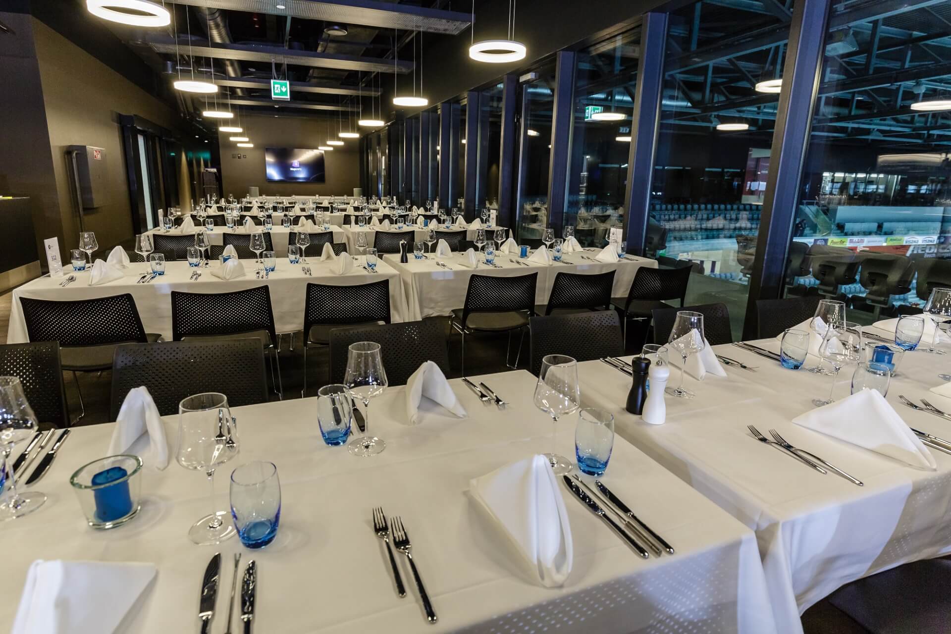 Ein moderner Essbereich mit weißen Tischtüchern, fein säuberlich angeordnetem Besteck, blauen Gläsern und gefalteten Servietten, der für eine Veranstaltung mit Tisch- und Stuhlreihen eingerichtet ist.