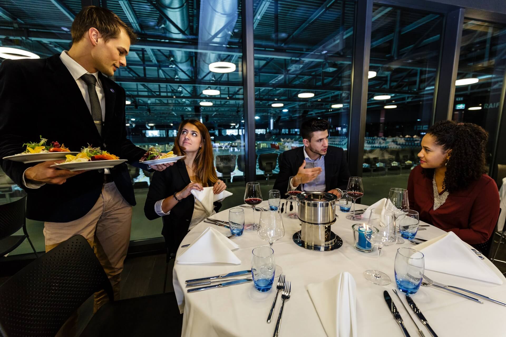 Ein Kellner serviert einer Gruppe von vier Personen, die an einem runden Tisch in einem modernen Restaurant Platz genommen haben, das Essen.