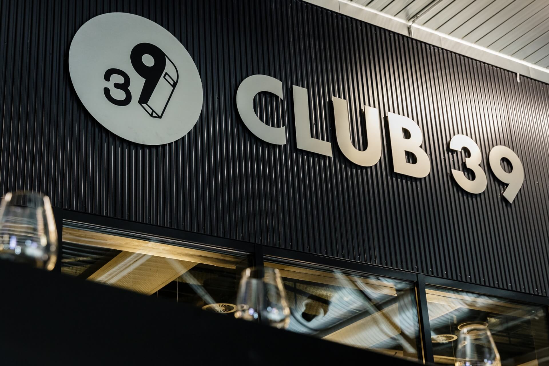 Schwarze Gebäudefassade mit großem CLUB 39-Schild und einem kreisförmigen Logo mit der Zahl 39. Im Vordergrund sind Glaswaren zu sehen.