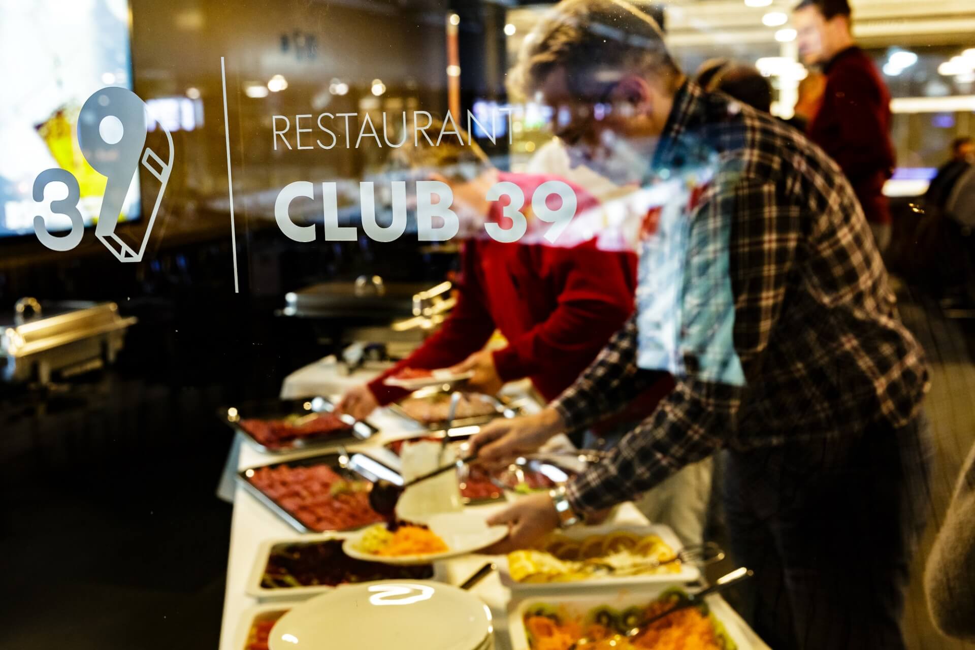 Menschen, die sich an einem Buffet im Restaurant Club 39 bedienen, gesehen durch ein Glasfenster mit dem Logo des Restaurants.