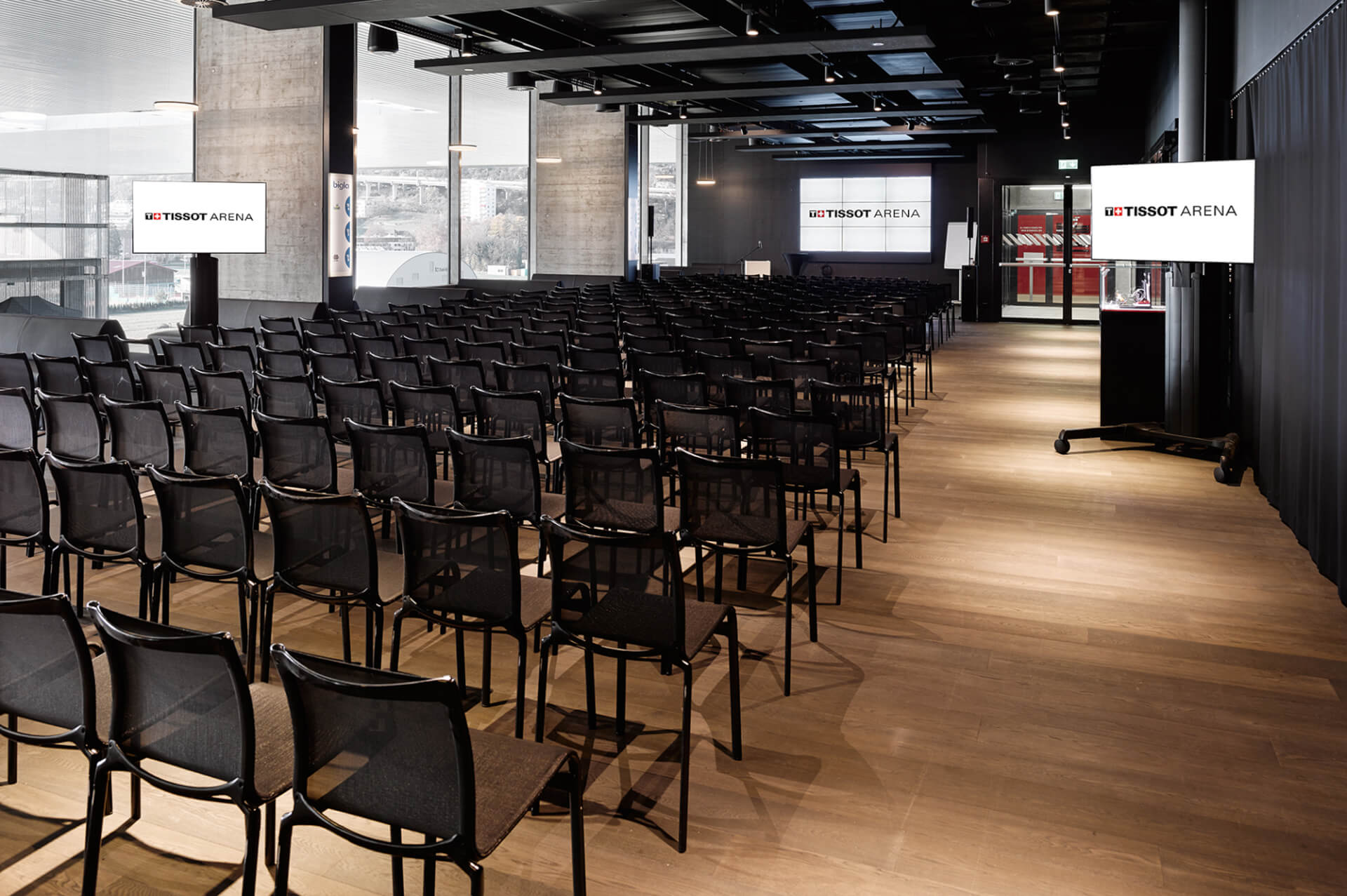 In einem modernen, gut beleuchteten Konferenzraum stehen leere schwarze Stühle vor zwei großen Bildschirmen mit dem Logo der Tissot Arena.