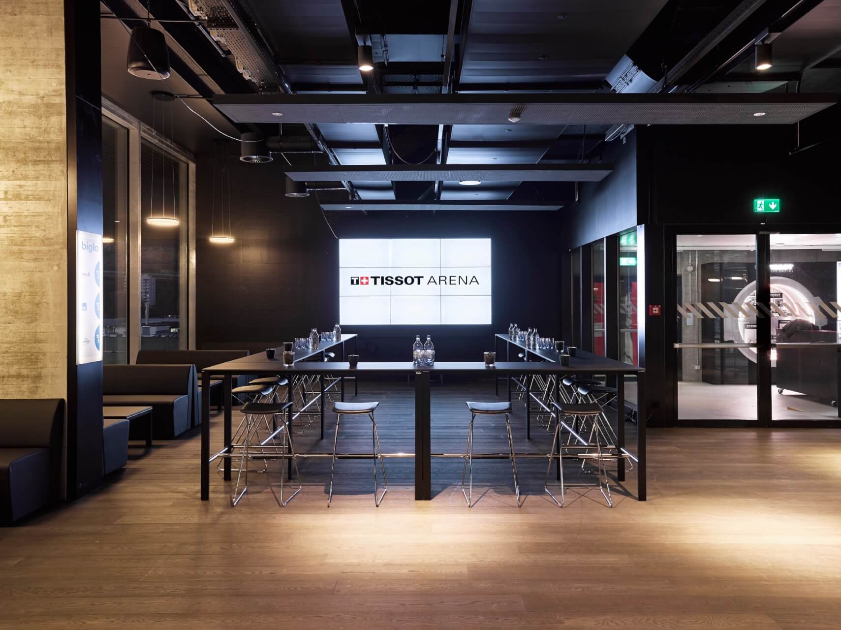 Ein moderner Konferenzraum mit U-förmig angeordneten Tischen, Wasserflaschen und einem großen Bildschirm mit der Tissot Arena an der Rückwand.