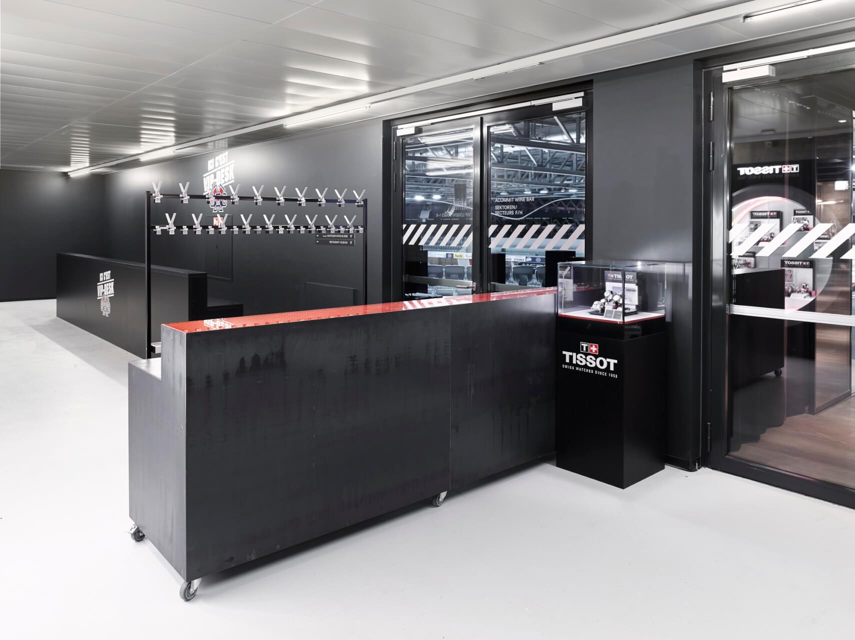 Ein moderner schwarzer Empfangstresen mit einer Tissot-Vitrine, eine Garderobe an der Wand und große Fenster, die ein industrielles Interieur zeigen.