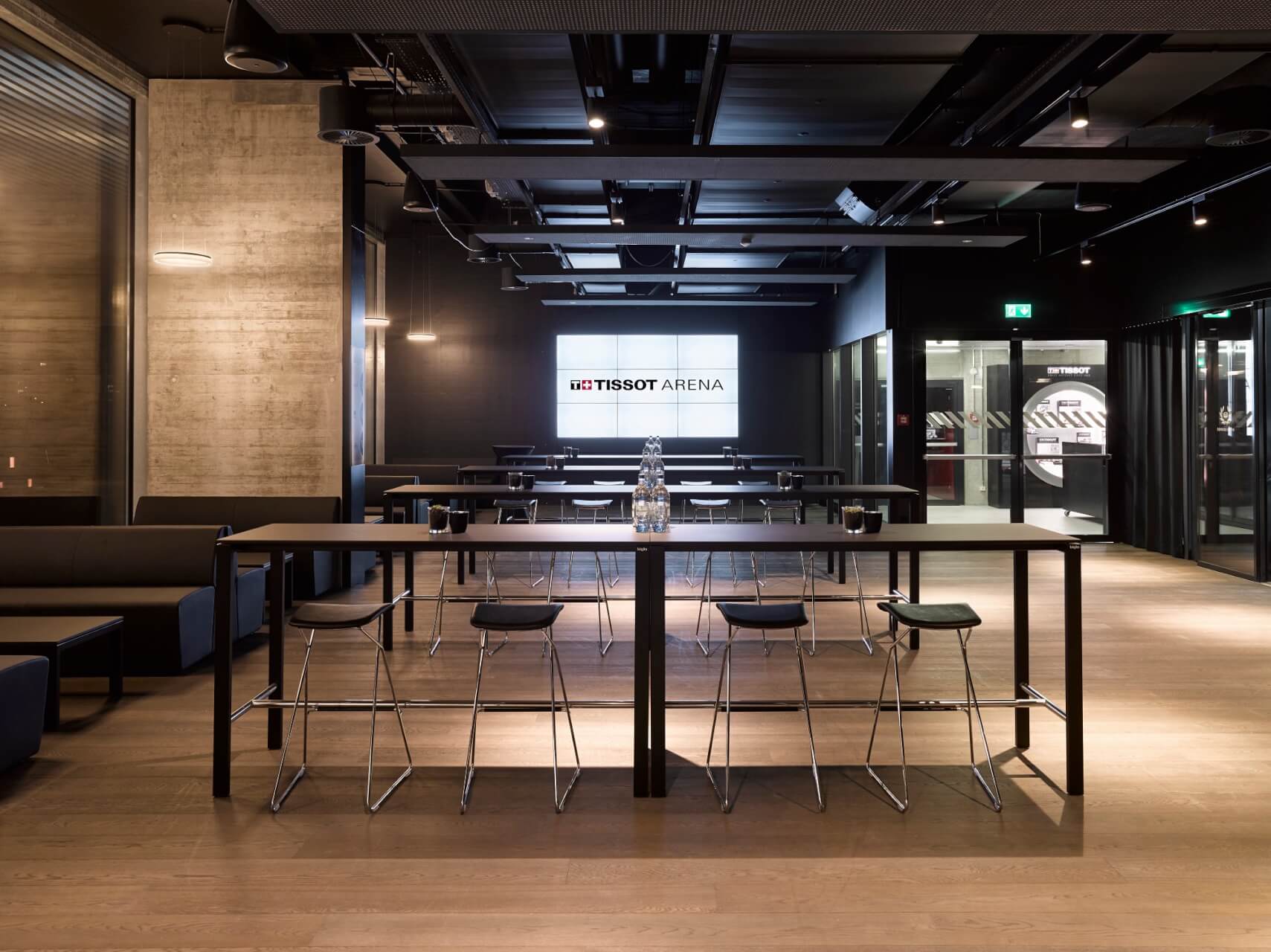 Moderner Lounge-Bereich mit Stehtischen, Barhockern und einem großen Bildschirm, auf dem die Tissot Arena gezeigt wird, in einem geräumigen, schummrigen Raum.