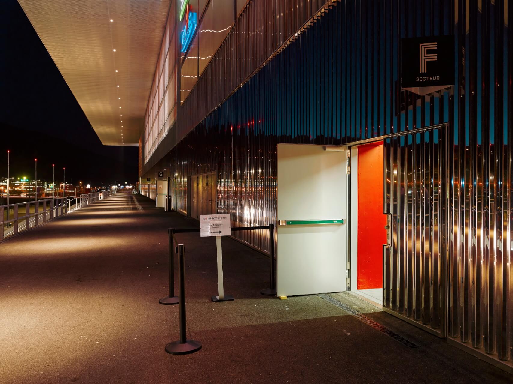 Eine hell erleuchtete, offene Tür mit roter Innenseite befindet sich in einer nächtlichen Metallfassade mit einem Gehweg und Pollern daneben.