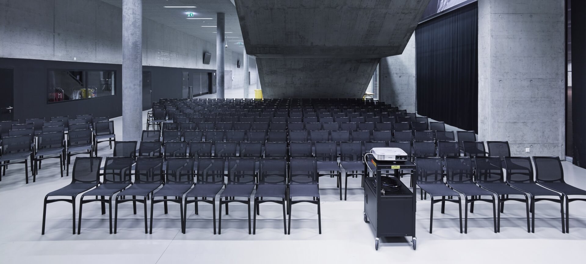 Reihen leerer schwarzer Stühle stehen vor einer Projektionsfläche in einem modernen, betonierten Auditorium, in dessen Vordergrund ein Projektor auf einem Wagen steht.