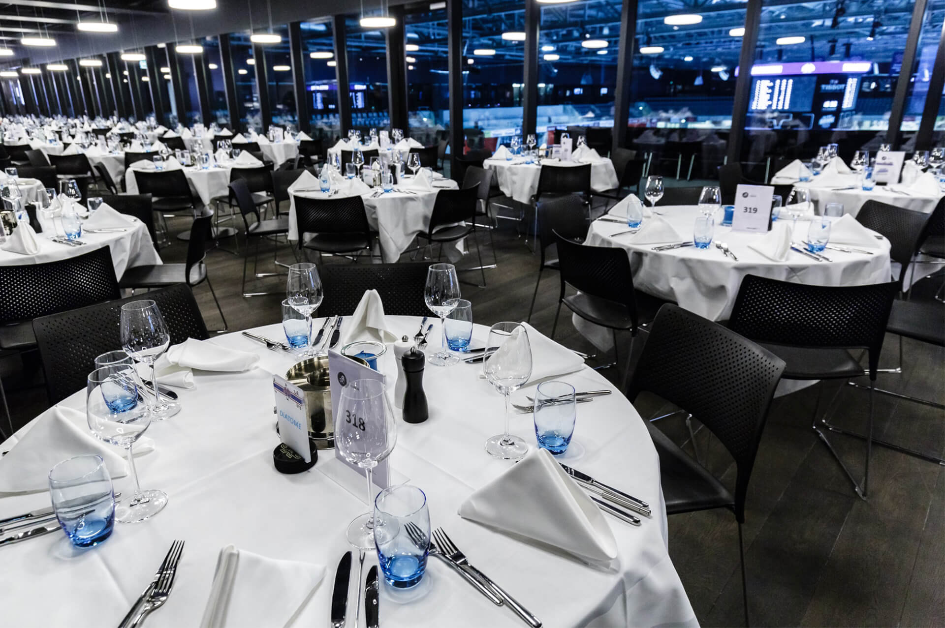 Großer Essbereich mit zahlreichen runden Tischen, die mit weißen Tischdecken, gefalteten Servietten, Gläsern und Besteck eingedeckt sind und durch große Fenster einen Blick auf eine Reithalle bieten.