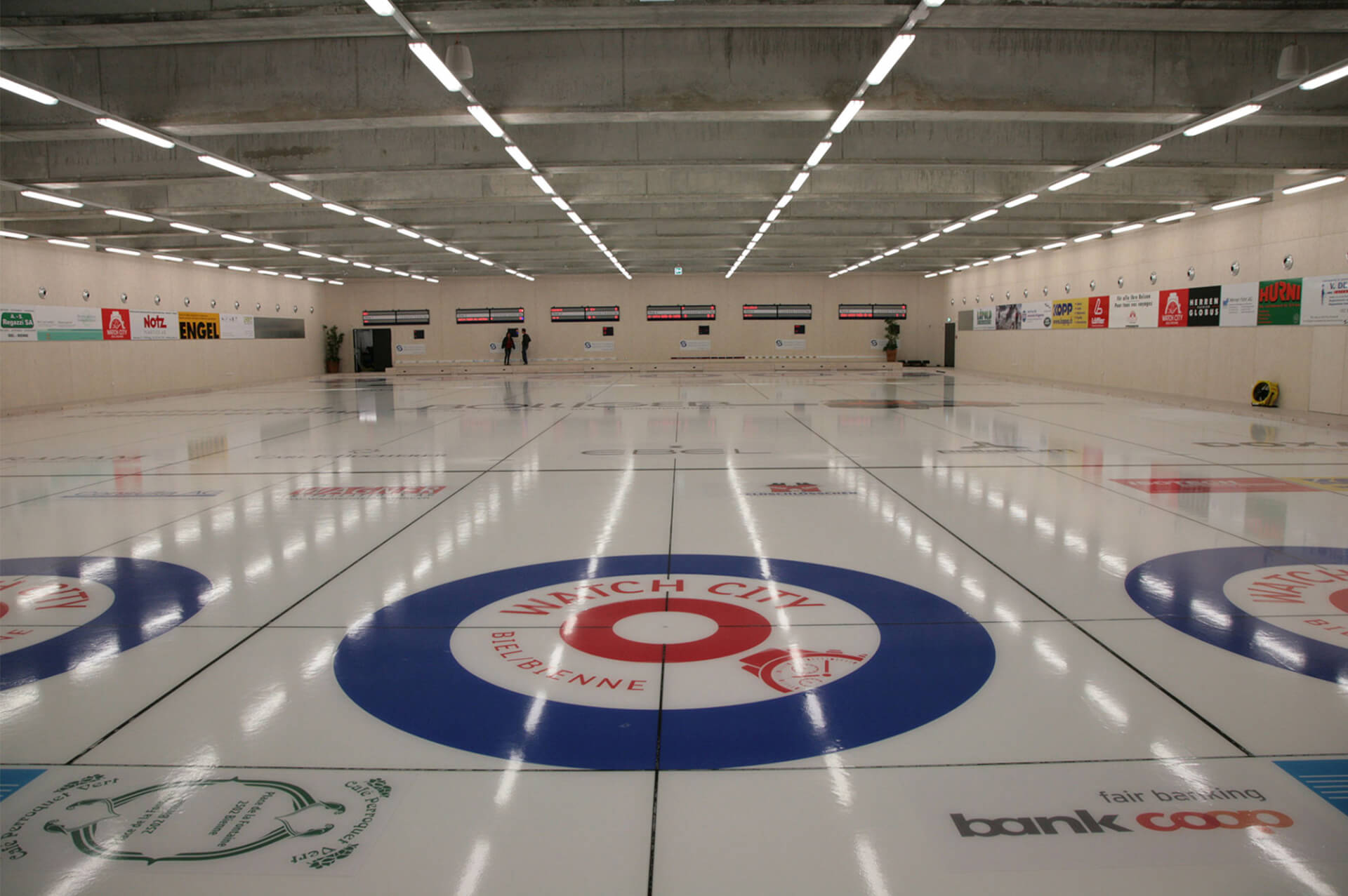 Indoor-Curlingbahn mit mehreren Curlingbahnen, Zielkreisen und verschiedenen Werbungen an den umliegenden Wänden.