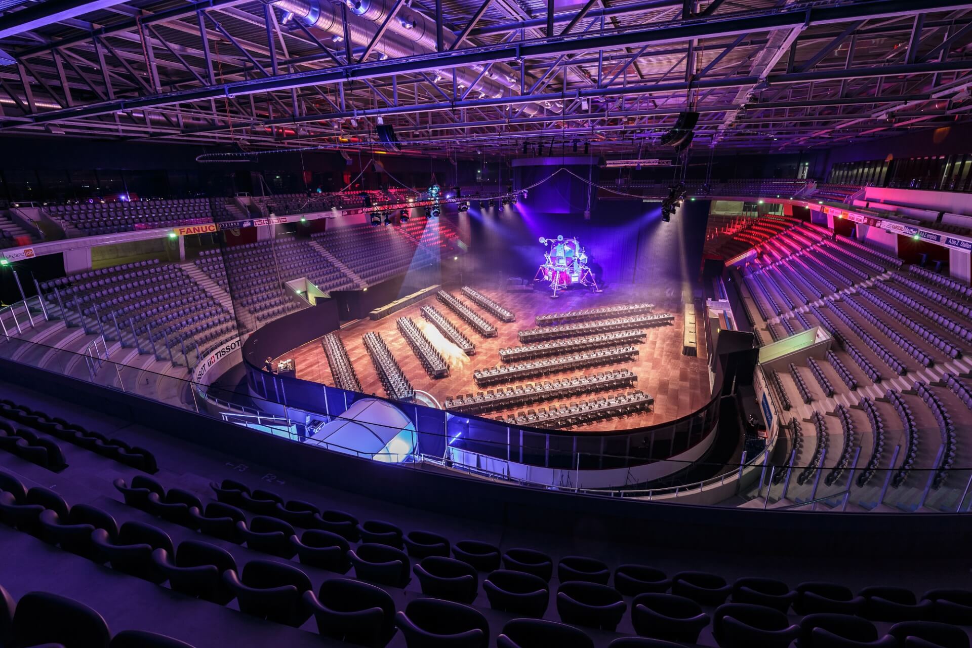 Große überdachte Arena mit leeren Sitzreihen vor einer Bühne, die von bunten Scheinwerfern beleuchtet wird, und wenig Publikum.