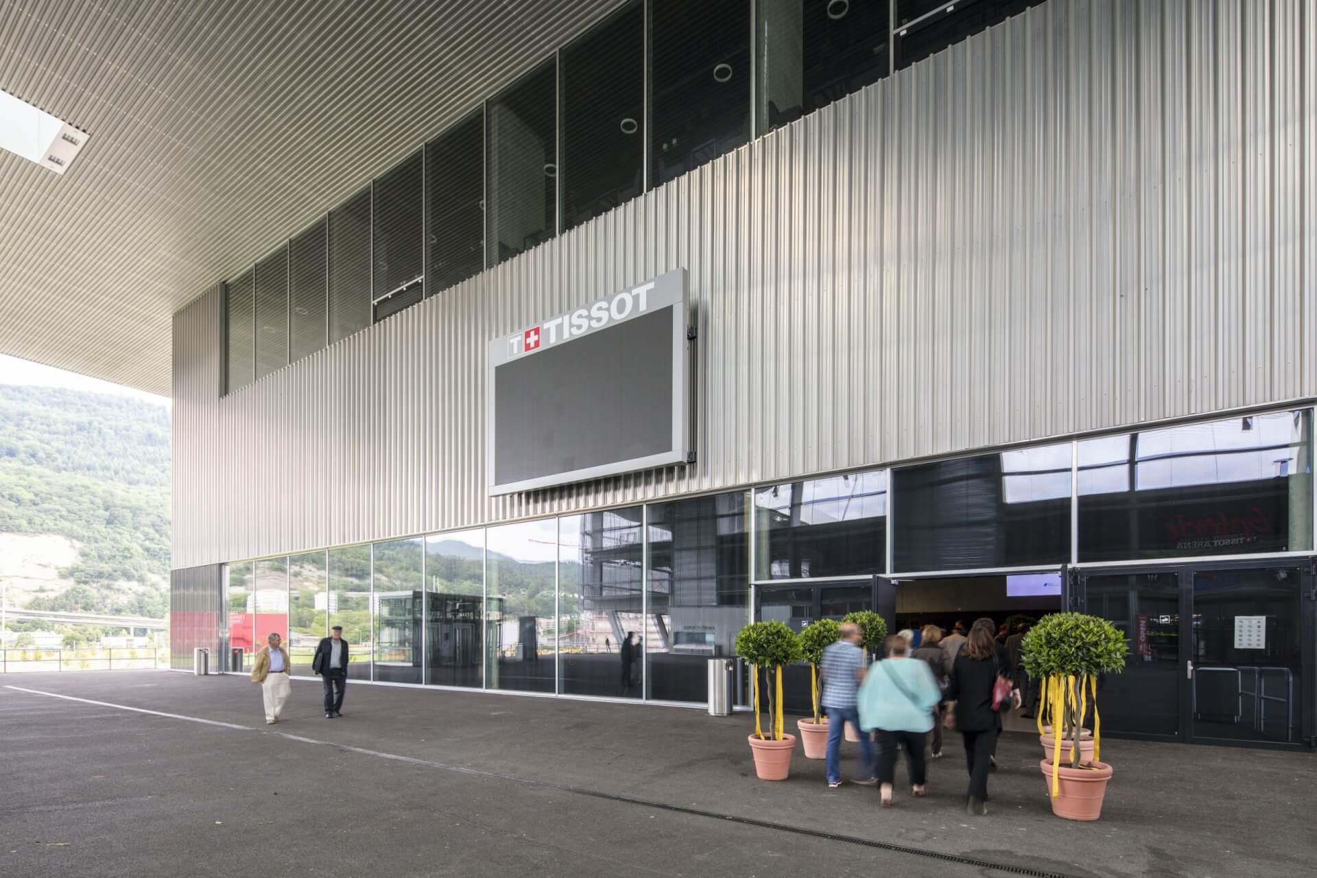 Menschen gehen auf den Eingang eines modernen Gebäudes zu, über dessen Türen ein großer leerer Bildschirm mit der Aufschrift TISSOT angebracht ist; Topfpflanzen säumen den Eingang.