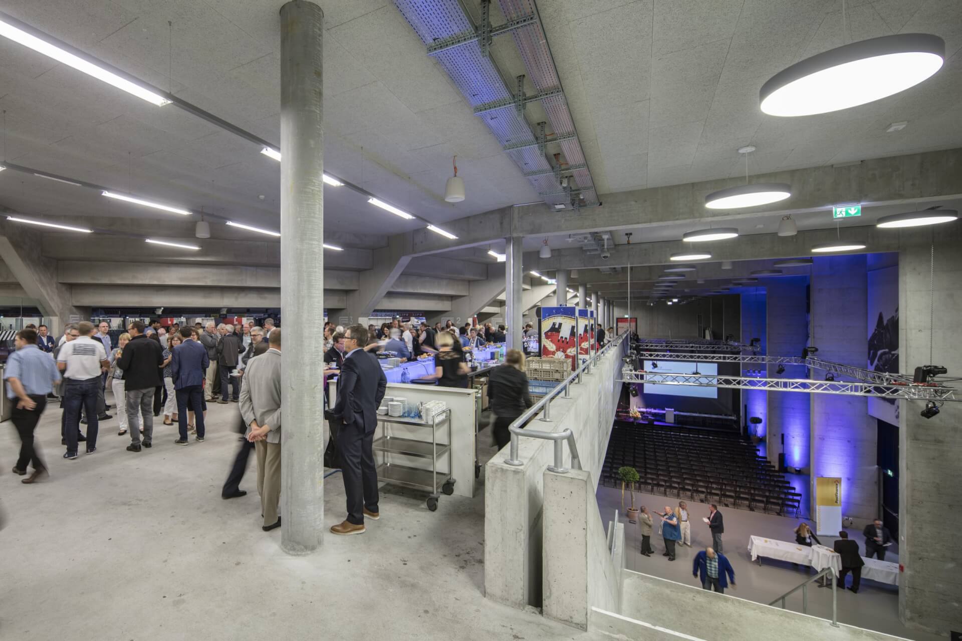 In einem modernen, betonierten Veranstaltungsraum mit einer oberen und einer unteren Ebene kommen Menschen zusammen und unterhalten sich; einige Tische und Ausstellungsstände sind sichtbar.
