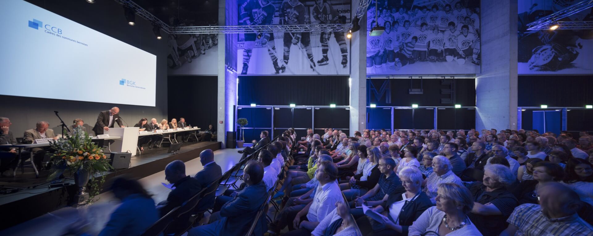 Ein großes Publikum nimmt an einer formellen Veranstaltung oder Sitzung in einem modernen Auditorium mit Sitzplätzen, Rednern auf der Bühne und großen Wandgemälden zum Thema Sport teil.