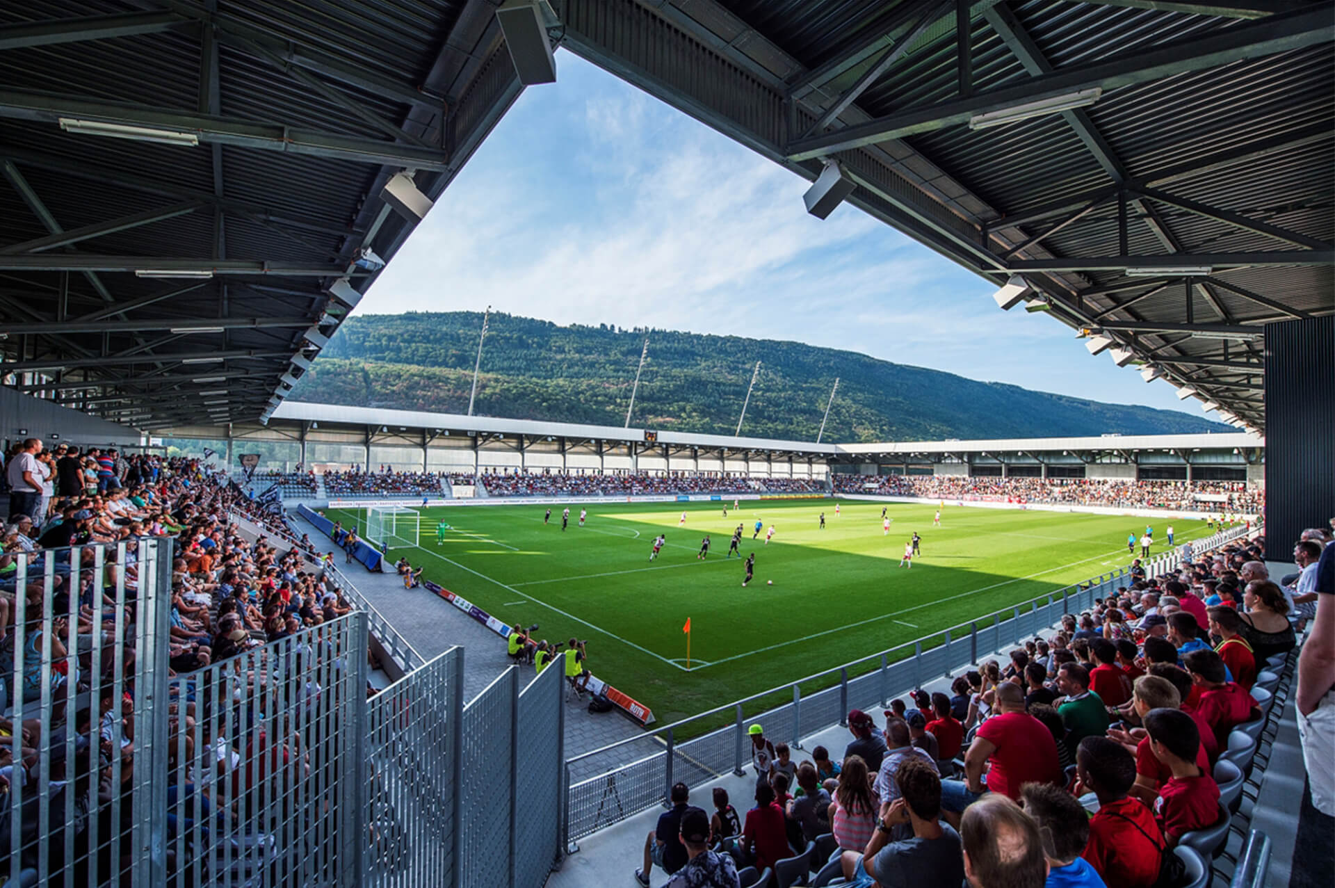 Ein Fußballspiel findet in einem modernen Stadion statt, das von Zuschauern umringt ist, vor einer Kulisse aus grünen Hügeln und blauem Himmel.