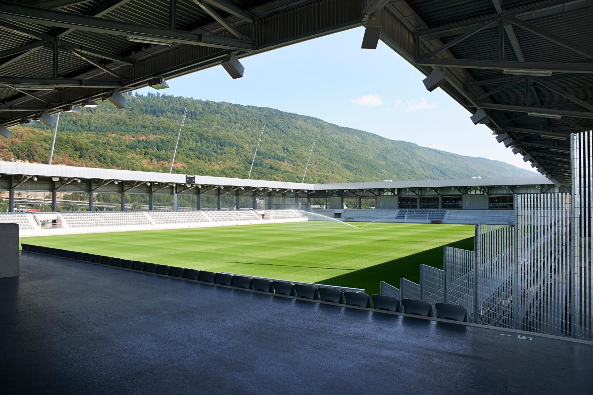 Ein modernes, leeres Fußballstadion mit grünem Rasen, überdachten Tribünen und den umliegenden Hügeln im Hintergrund.