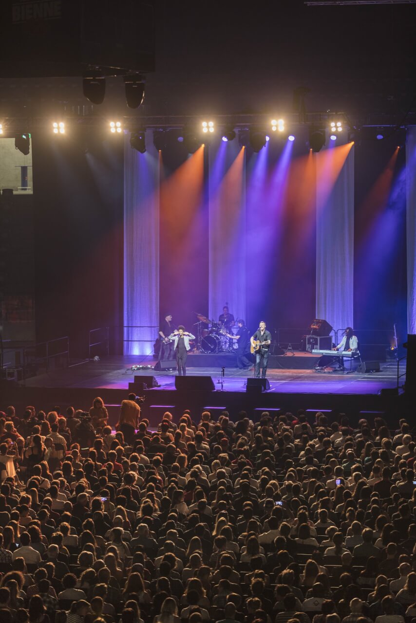 Eine Band tritt auf einer hell erleuchteten Bühne mit bunten Lichtern auf, während ein großes Publikum im Vordergrund zusieht.