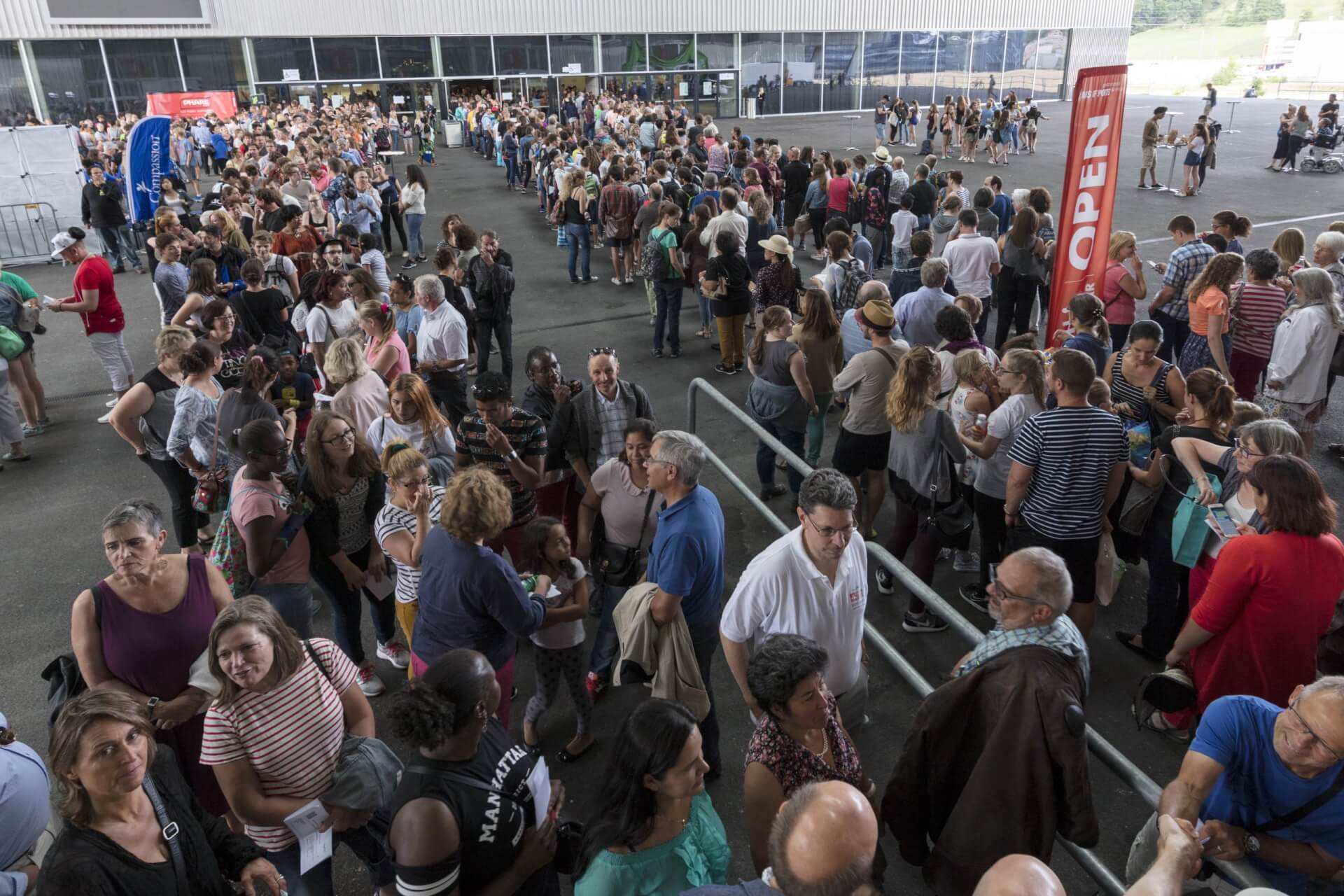 Eine große Menschenmenge steht in einer Schlange vor einem Gebäude, in der Nähe von Absperrungen und einem OPEN-Schild, und wartet darauf, hineinzukommen.