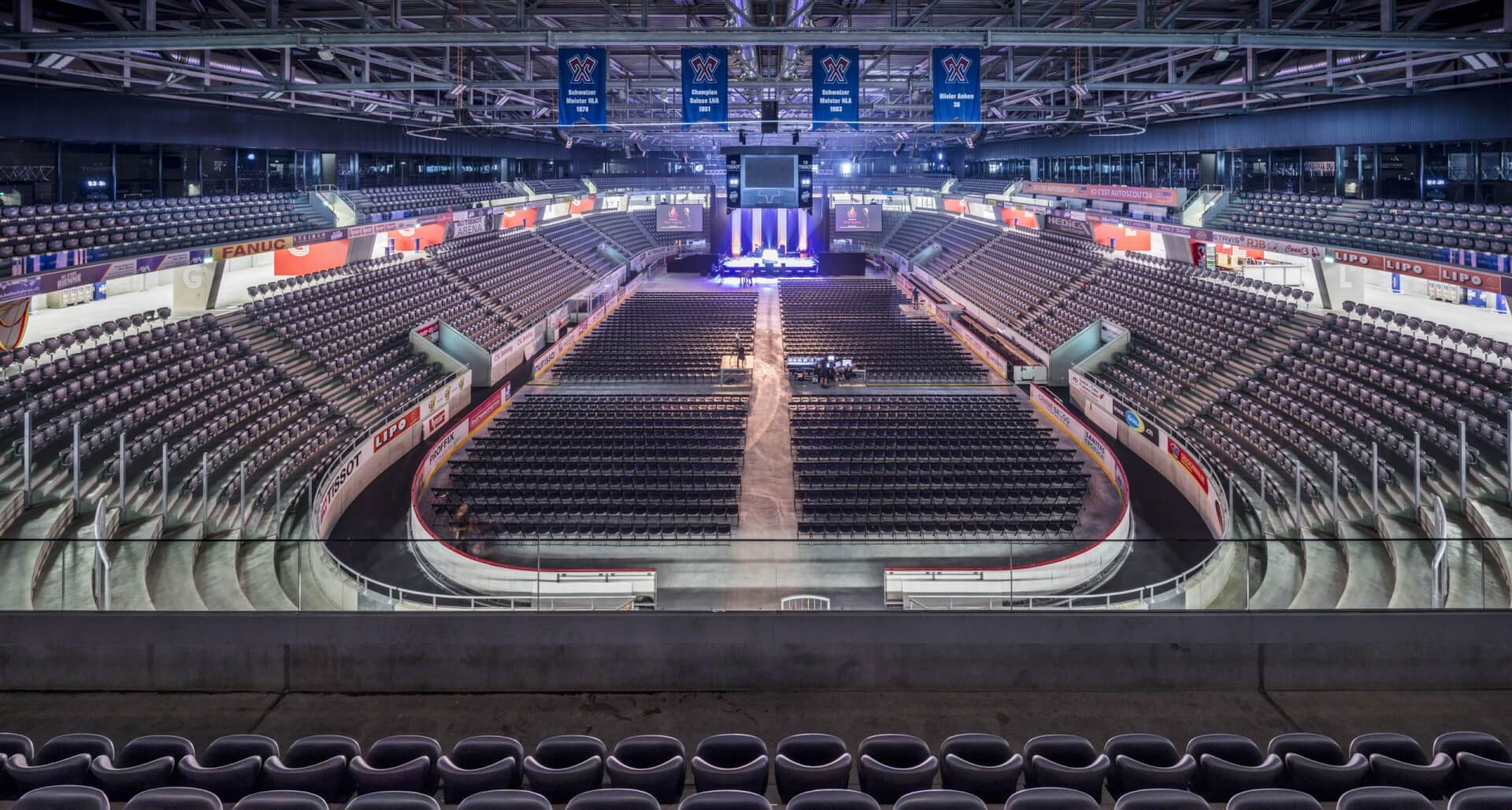 Leere Halle mit Sitzreihen, einer großen zentralen Bühne mit Beleuchtung und mehreren von der Decke hängenden Bannern.