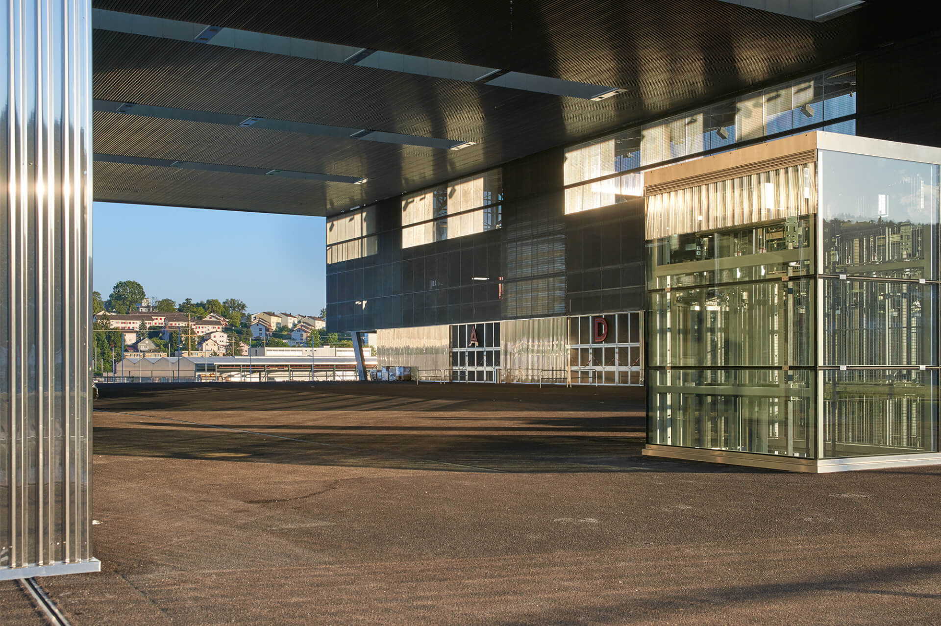 Modernes Gebäude mit reflektierendem Glasaufzug, Wellblechwand und offener gepflasterter Fläche im Sonnenlicht; im Hintergrund sind entfernte Häuser zu sehen.